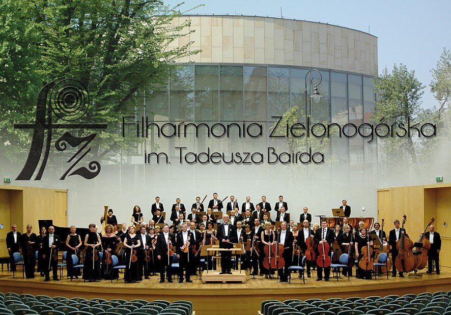 Orkiestra symfoniczna stojąca na scenie sali koncertowej Filharmonii Zielonogórskiej im. Tadeusza Bairda. Muzycy z instrumentami smyczkowymi, dętymi i perkusyjnymi ustawieni są w kilku rzędach, a za nimi na ścianie widoczny jest duży napis z nazwą filharmonii oraz jej logo.