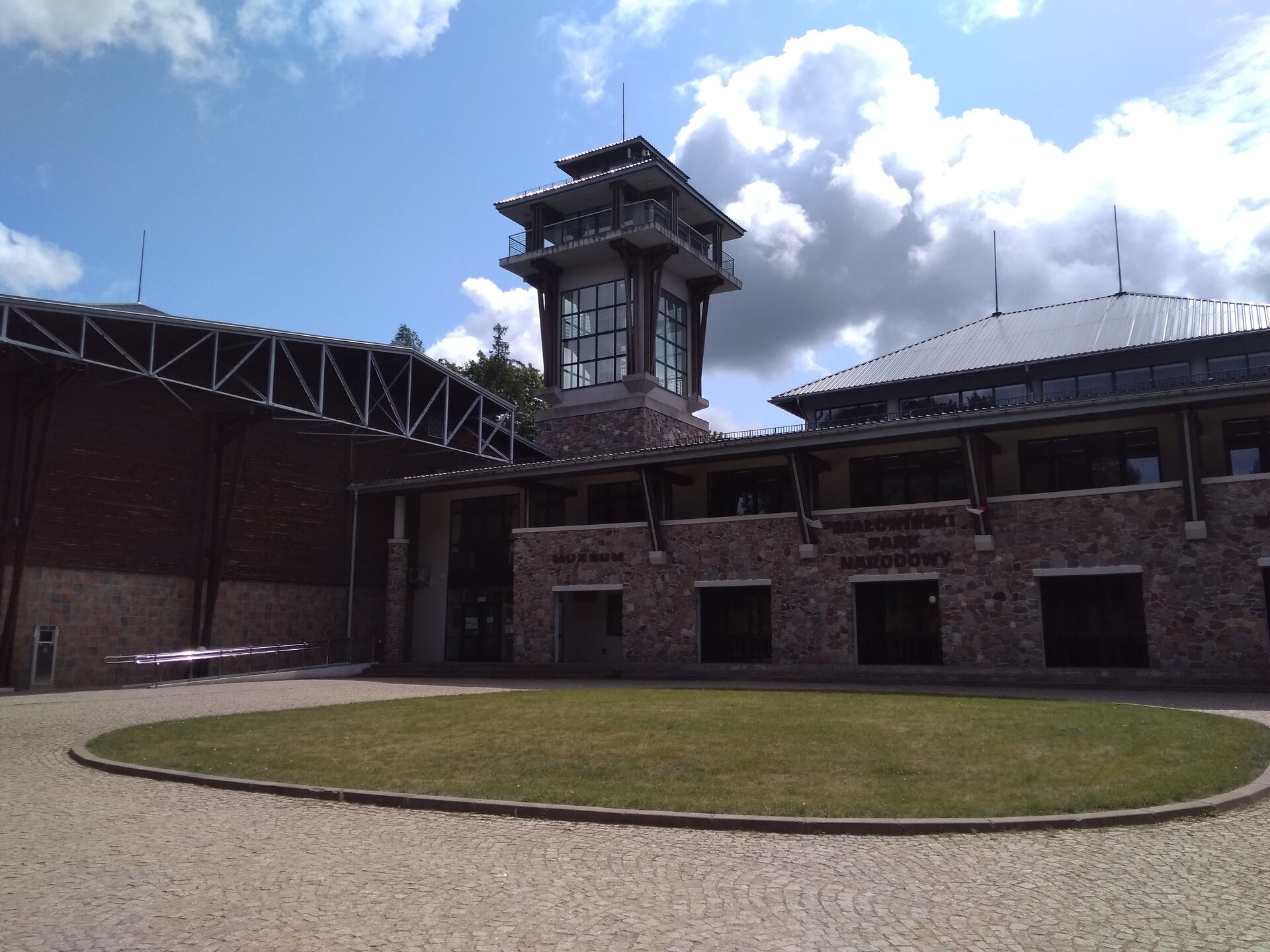 Fotografia przedstawia budynki Muzeum Przyrodniczo‑Leśnego w Białowieskim Parku Narodowym składające się z podłużnego piętrowego budynku oraz przeszklonej wieży z tarasem widokowym. Przed budynkami znajduje się brukowany plac z okrągłą trawiastą częścią w centralnej części.