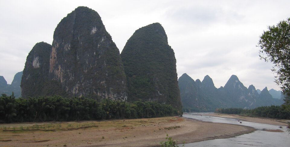 Ilustracja przedstawia wzgórza mogotów w okolicy Guilin  w Chinach. Wysokie, podłużne skały górują nad biegnącą dołem rzeką. U ich stóp rośnie niski las.