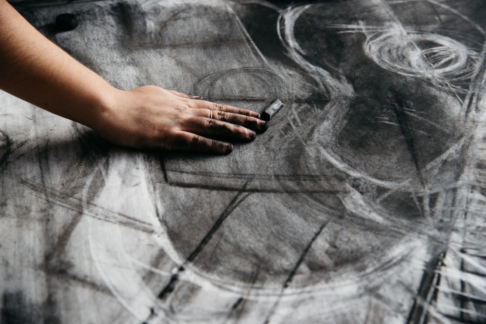 Zdjęcie przedstawia zbliżenie na rysunek wykonywany węglem. Dużych rozmiarów karton pokryty jest w różnych kierunkach prostymi kreskami, częściowo roztartymi oraz kolistymi przecierkami wykonanymi na rysunku gumką. Ręka autora, z ubrudzonymi węglem palcami opiera się o kartkę, obok dłoni leży pałeczka węgla rysunkowego.