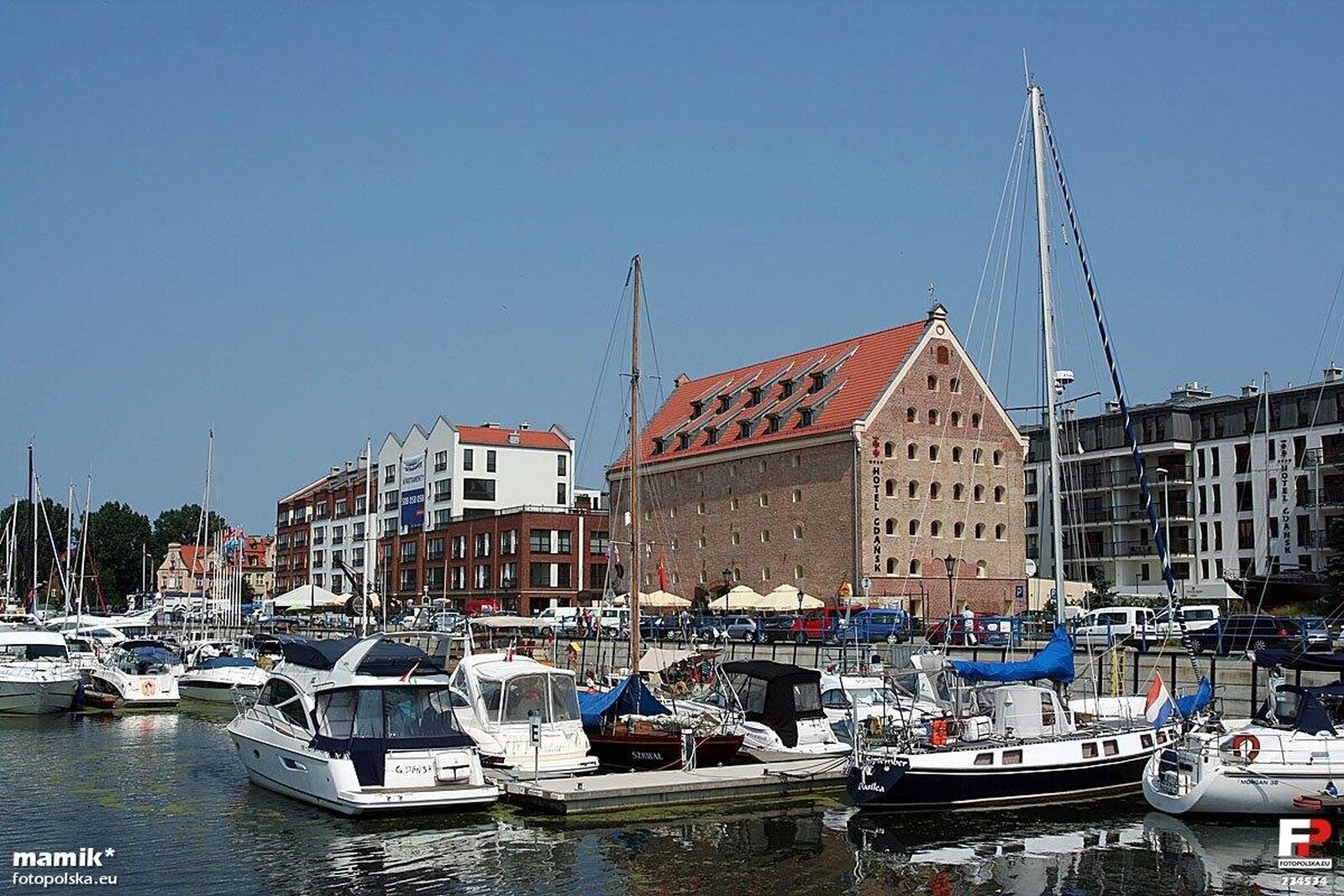 Fotografia przedstawia jachty zacumowane w niewielkiej przystani. Są niewielkie, mają biały kolor. W tle zabudowania miejskie.
