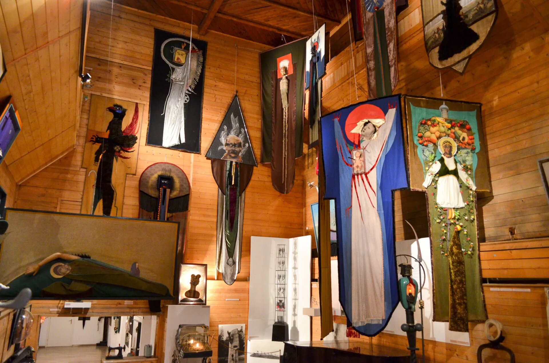 Zdjęcie przedstawia wnętrze galerii artystycznej. Na ścianach wiszą obrazy. Z sufitu zwisają dzieła na płótnie. W pracach przewijają się motywy religijne, przedstawienia postaci w aureolach, jedna otoczona owocami, druga  zdrewnianą, trójwymiarową ręką. Na ścianie wisi wizerunek istoty podobnej do czarnego ptaka. Dzieła składają się z różnych materiałów i mają nieregularne kształty.