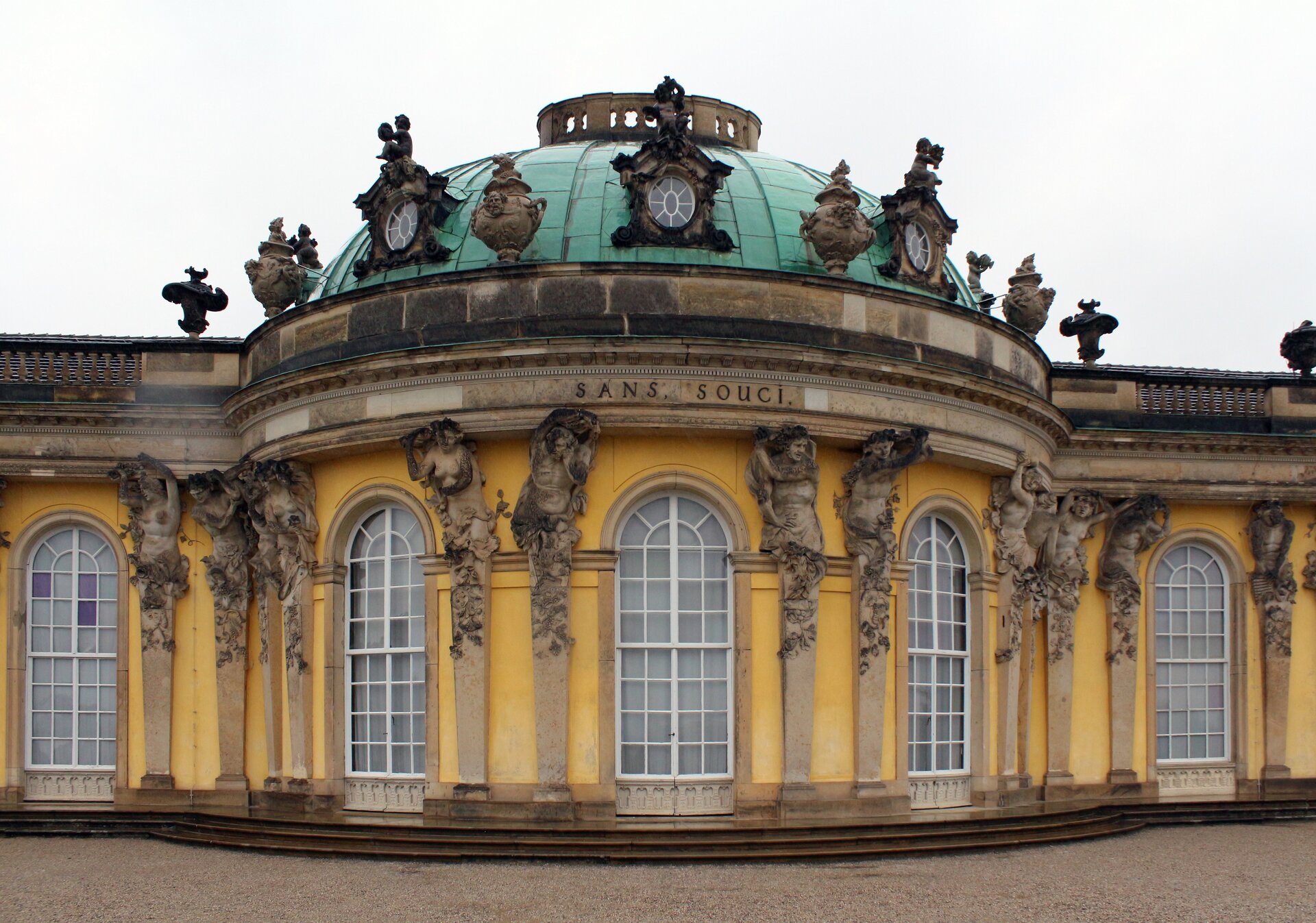 Ilustracja przedstawia zbliżenie na fragment elewacji południowej Pałacu Sanssousi w Poczdamie. Hermy półokrągłego ryzalitu zwieńczone są postaciami z orszaku Bachusa, sprawiającymi wrażenie, że podtrzymują balustradę wieńczącą kondygnację. Na balustradzie znajdują się rytmicznie rozmieszczone wazony. Na kopule, tuż nad balustradą są umieszczone niewielkie, okrągłe okna zwieńczone aniołkami.