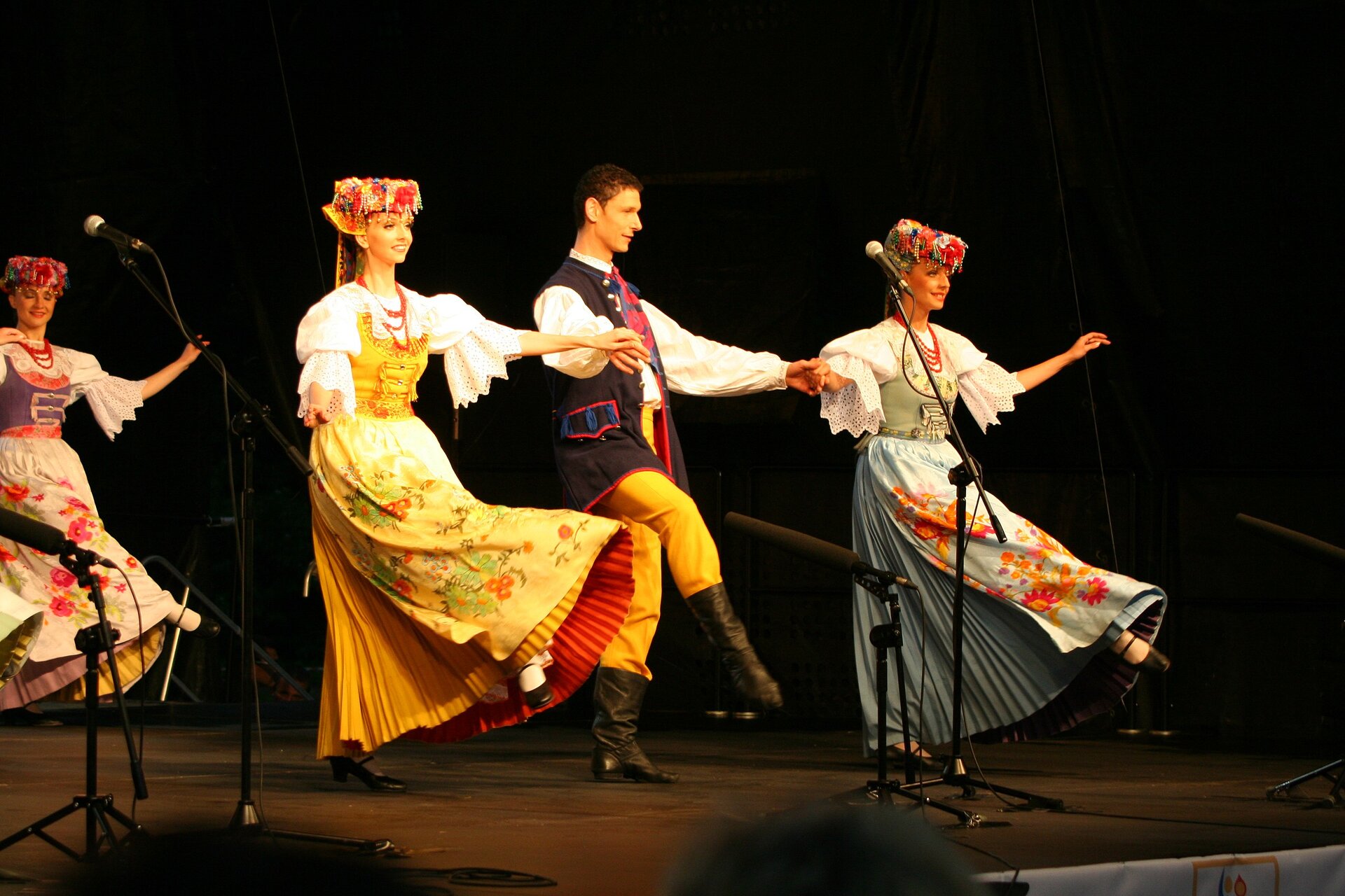 Fotografia tańczących osób w strojach ludowych. Na pierwszym planie mężczyzna, trzyma za ręce tańczące po bokach kobiety. Ubrani w tradycyjne, kolorowe stroje. Kobiety mają długie suknie z falbanami, na głowie mają czepce z kwiatów. Mężczyzna nosi czarne buty do kolan, pomarańczowe spodnie, białą koszulę i czarną zdobioną kamizelkę. W tle kolejna tancerka.