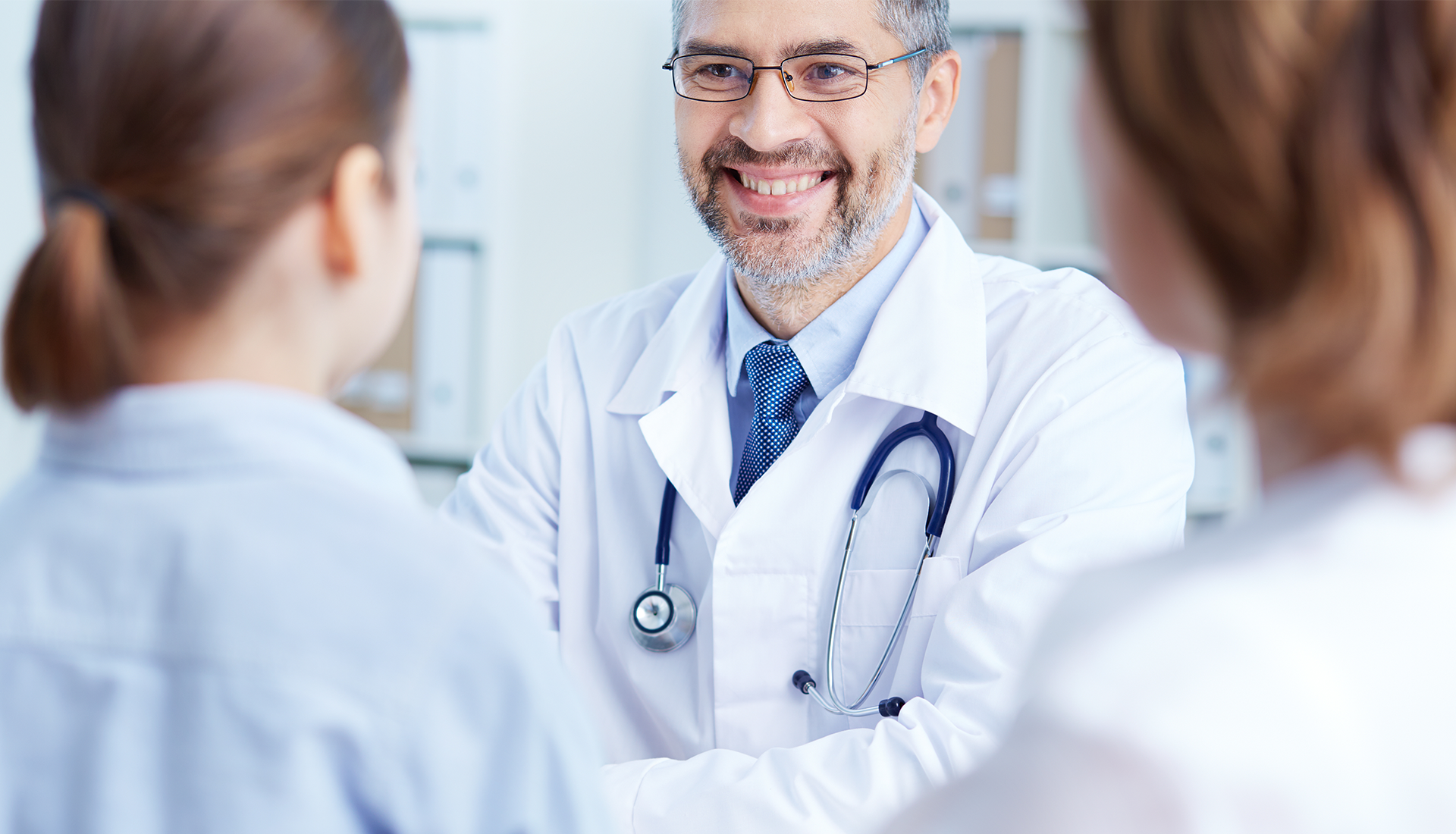 Zdjęcie przedstawia młodego, uśmiechniętego mężczyznę w białym fartuchu lekarskim, na szyi przewieszony ma stetoskop lekarski. Pochłonięty jest rozmową z dwiema osobami w białych fartuchach.