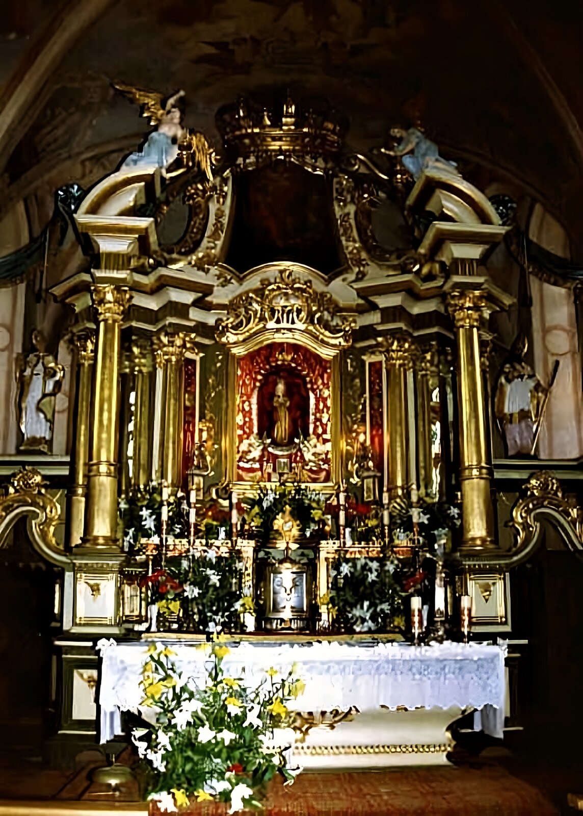 Zdjęcie przedstawia ołtarz, który jest bogato zdobiony złotem. W jego centralnej części znajduje się sylwetka kobiety w długiej, złotej szacie. Wokół niej widoczne są złote kolumny. Nad ołtarzem widoczne są dwa anioły w niebieskich szatach i złotych skrzydłach. Przed ołtarzem znajduje się stół, na którym stoją wysokie, białe świece, kolorowe kwiaty, a w centralnej części krzyż i tabernakulum. Przed stołem znajduje się duży bukiet żółtych i białych kwiatów. 