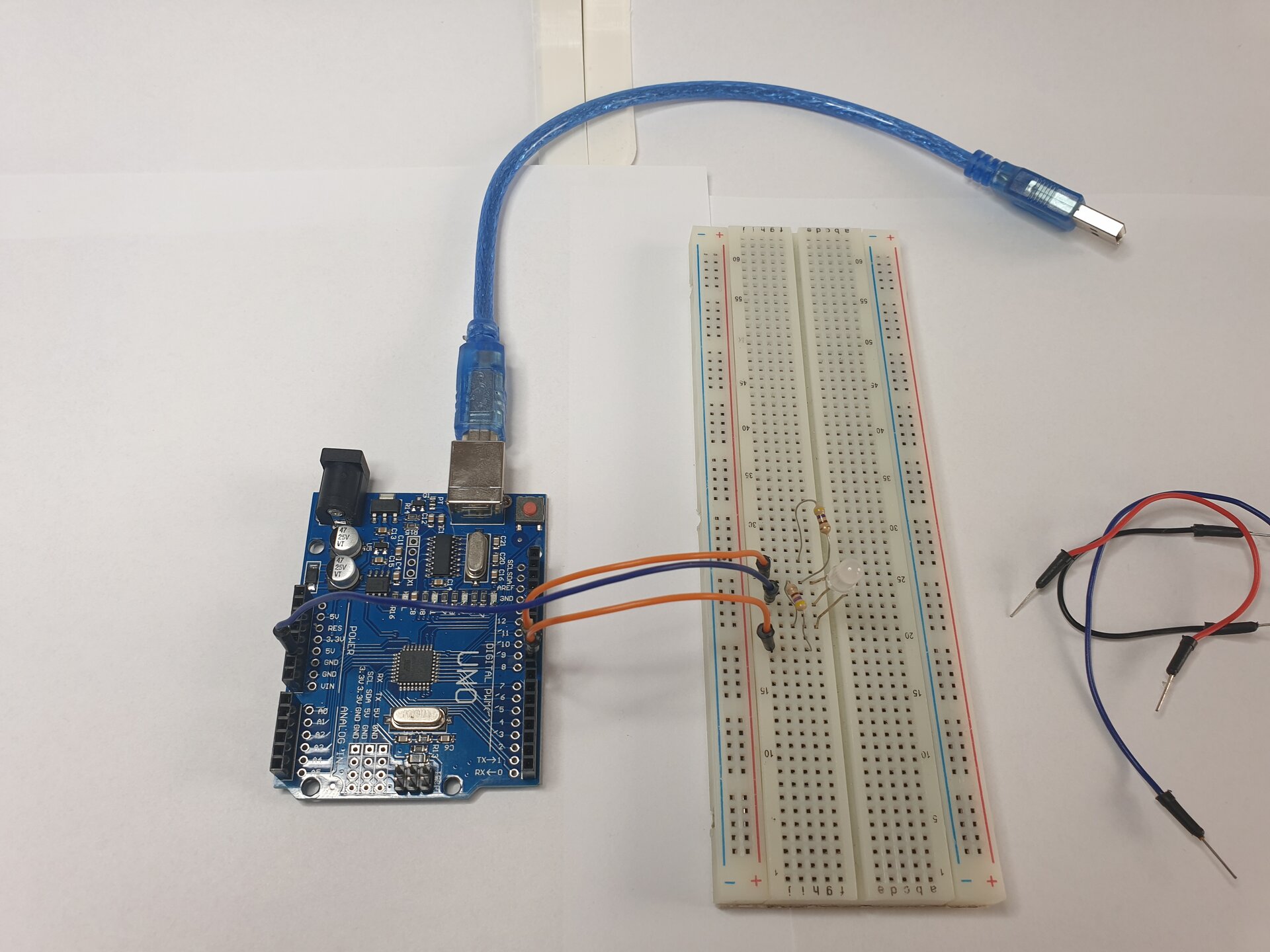 Zdjęcie przedstawia moduł Arduino z podłączonym do niego kablem USB, płytkę stykową, w którą wpięto diodę oraz leżące obok płytki przewody. Połączono moduł z płytką za pomocą przewodu wpiętego w module w 5 V i w płytce w tym samym wierszu, co środkowa nóżka diody. Dwa rezystory wpięto w płytkę stykową w taki sposób, że jedna nóżka każdego rezystora jest w tym samym wierszu, co zewnętrzne nóżki diody, a druga nóżka każdego rezystora jest wpięta „na zewnątrz” nóżek diody. Dwoma przewodami połączono nóżki diody z modułem Arduino. Pierwszy przewód wpięty jest w płytce w tym samym wierszu, co nóżka rezystora i nóżka diody, a w module wpięty jest do pinu D10. Drugi przewód wpięty jest w wierszu, w którym drugi rezystor ma nóżkę poza wierszem, w którym jest wpięta trzecia nóżka diody, a w module wpięty jest do pinu D9.