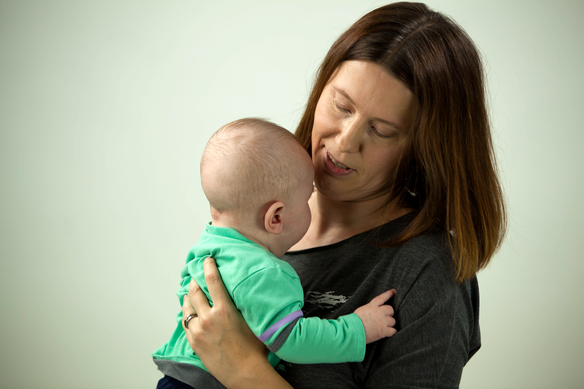 Fotografia przedstawia mamę trzymającą blisko siebie niemowlę. Niemowle zwrócone jest tyłem do odbiorcy ilustracji, mama przodem. Ilustracja pokazuje więź pomiędzy matką, a dzieckiem tworzącą się min poprzez poznawanie naturalnych zapachów bliskiej osoby.