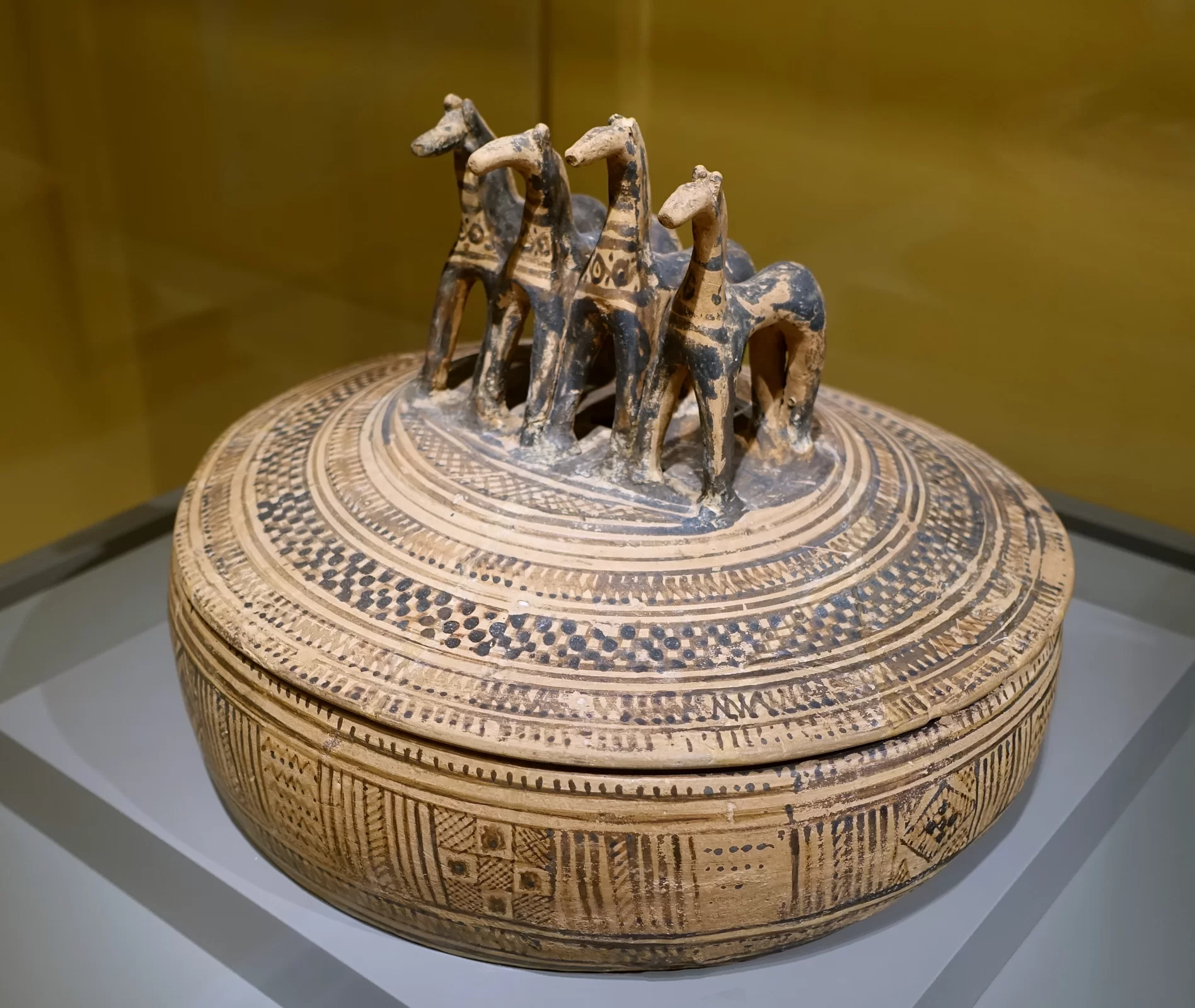 Ilustracja przedstawia ceramiczne naczynie z pokrywką. Naczynie ma okrągły, wypukły korpus i niską, kopulastą pokrywkę. Na wieku ustawione są cztery konie. Wokół koni wieko zdobione jest ornamentami ułożonymi w pasy. Na bocznych ściankach naczynia znajdują się geometryczne wzory.