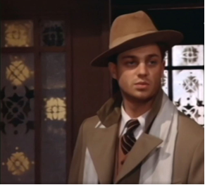 Zdjęcie przedstawia młodego mężczyznę. Ma pociągłą twarz i lekko zarysowany zarost. Ma na głowie brązowy kapelusz. Ubrany jest w garnitur i brązowy płaszcz z białym szalem.