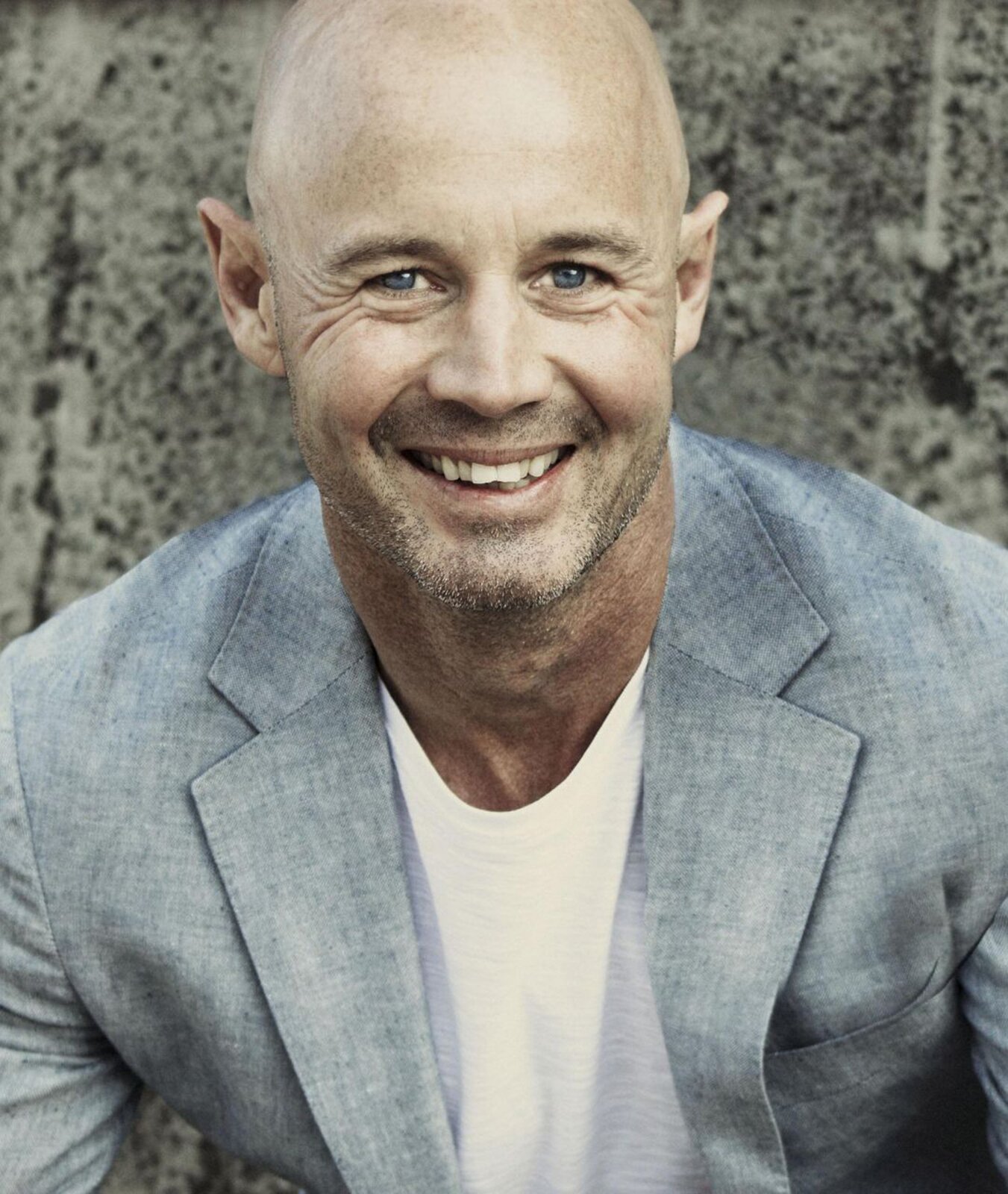 Zdjęcie przedstawia uśmiechniętego, łysego mężczyznę w średnim wieku, ubranego w białą koszulkę i szarą marynarkę.