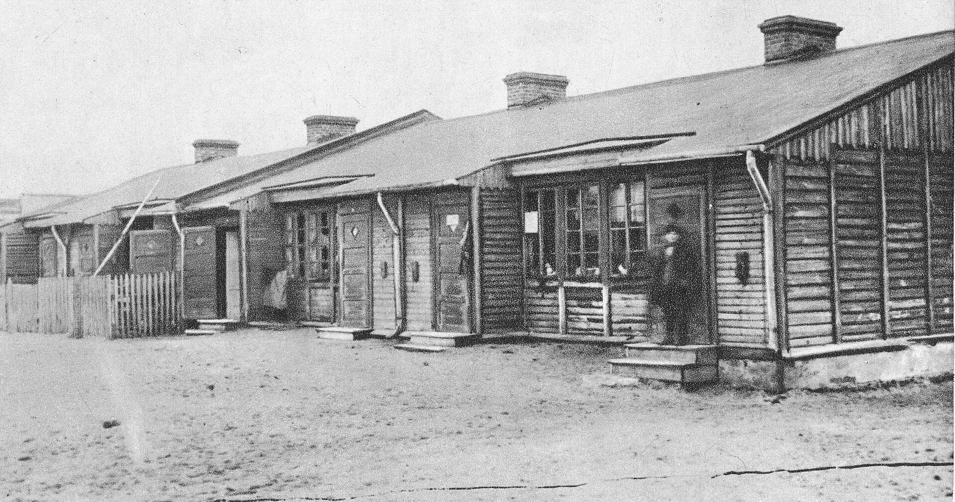 Zdjęcie przedstawia dwa drewniane baraki. Są one ustawione szeregowo, jeden obok drugiego. Baraki są parterowe. Mają lekko spadziste dachy. Przed wejściem do drugiego baraku stoi drewniany płot.