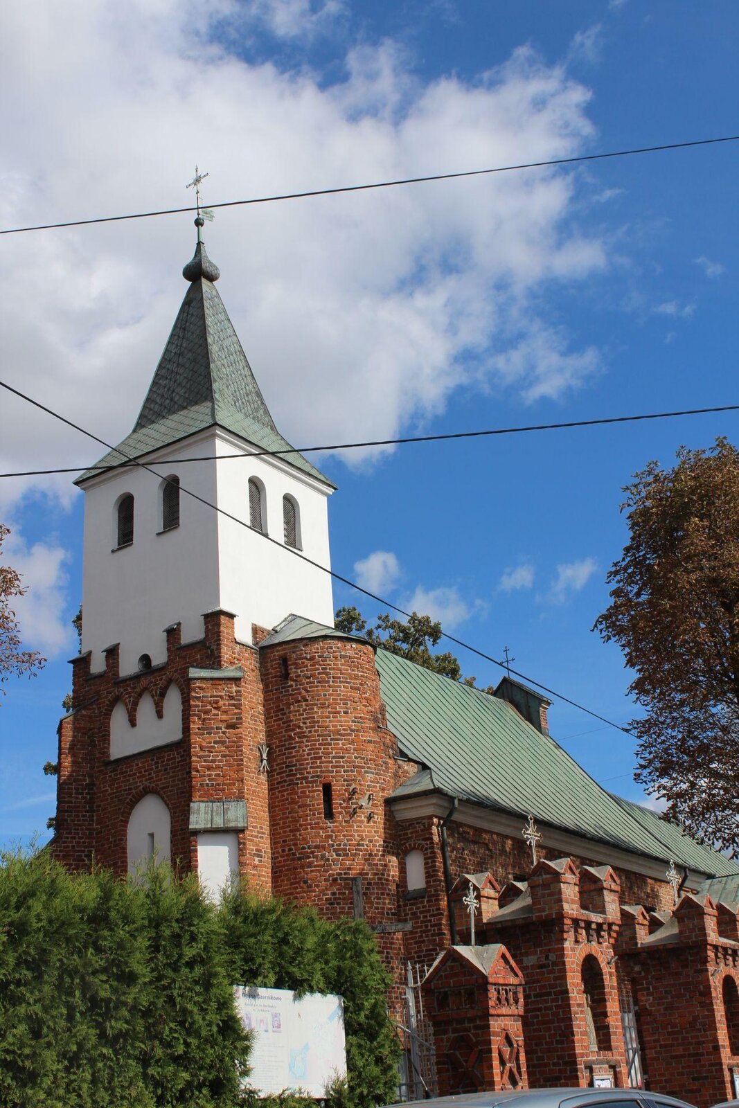 Zdjęcie przedstawia bok kościoła. Widoczna jest wieża, umieszczona na przodzie kościoła, która wykonana jest częściowo z czerwonej cegły, a częściowo z białego tynku. Na szczycie wieży skośny dach i krzyż. Za wieżą widoczna jest nawa główna ze skośnym dachem. Nawa wykonana jest z czerwonej cegły. Przed kościołem zielone krzewy, przy których znajduje się wyblakła tablica informacyjna.