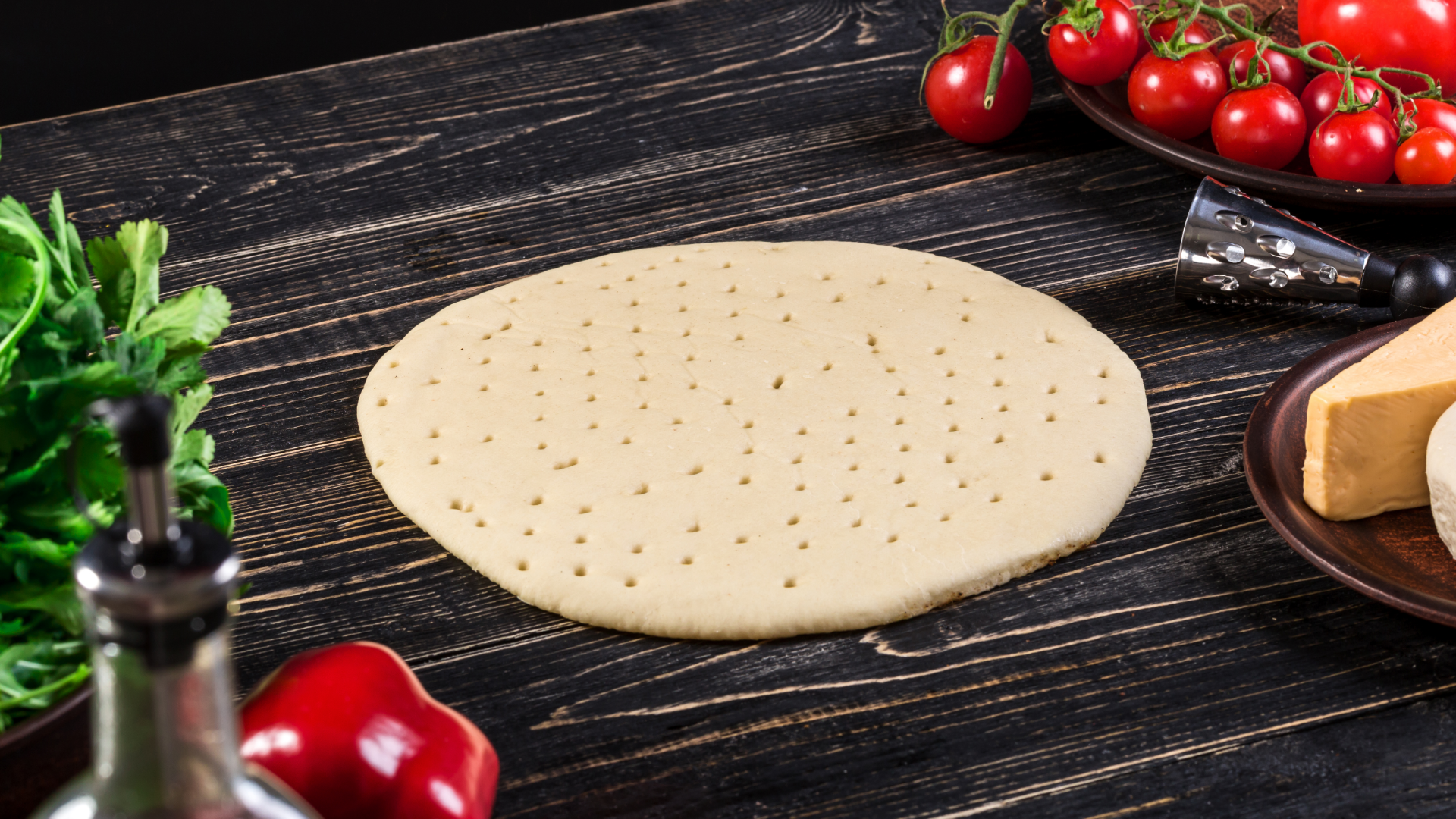 Zdjęcie przedstawia spód do pizzy, który leży na drewnianym blacie. Obok na talerzach znajdują się produkty na pizzę. Widoczne są pomidorki koktajlowe, dojrzewający ser, papryka, liście oraz fragment butelki z oliwą.