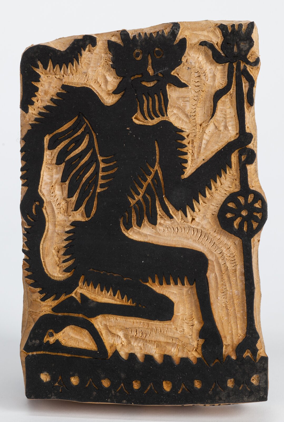 Zdjęcie przedstawia klocek drzeworytniczy, na którym wyrzeźbiono postać diabła. Jest to stwór pokryty futrem, o sylwetce przypominającej człowieka, ale z kopytami i długim ogonem oraz rogami na głowie. W ręce trzyma wysoki kij z okrągłym elementem na środku. Wszystkie wypukłe elementy są pomalowane na czarno.