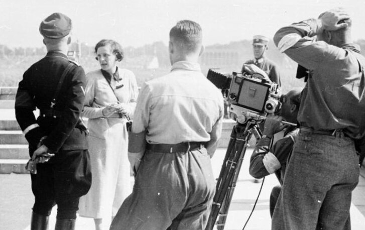 Fotografia wykonana jest w plenerze. Przedstawia Leni Riefentstahl i pięciu mężczyzn, którzy stoją wokół kobiety i patrzą w jej kierunku. Jeden z mężczyzn jest operatorem kamery, która skierowana jest w kierunku reżyserki. Pierwszy mężczyzna z lewej to Heinrich Himmler. Zdjęcie pokazuje sylwetki mężczyzn od tyłu.