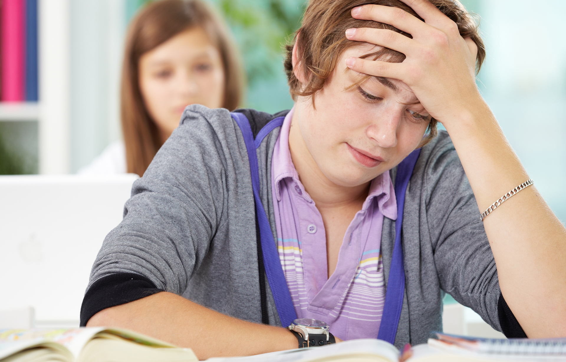 Zdjęcie przedstawia na pierwszym planie nastolatka siedzącego w przy blacie biurka. Chłopiec ma krótkie, jasne włosy, jest ubrany w fioletową koszulkę polo w paski i szarą, rozpinaną bluzę z fioletowymi troczkami. Łokieć lewej ręki opiera o blat, a otwartą dłonią dotyka czoła. Prawe przedramię oparte jest na blacie. Przed chłopakiem widoczne są otwarte książki. W tle widoczna rozmyta postać dziewczyny siedzącej przed otwartym laptopem.
