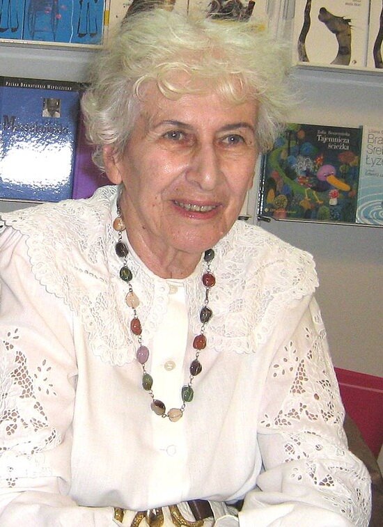 Kolorowa fotografia przedstawia portret uśmiechniętej starszej pani o krótkich siwych włosach. Ubrana jest w koronkową białą bluzkę, którą zdobi naszyjnik z kolorowych kamieni szlachetnych. W tle widać okładki książek dla dzieci, których jest autorką.