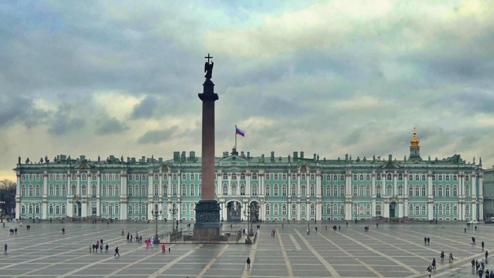 Zdjęcie przedstawia długi budynek w niebieskim kolorze z licznymi oknami, które posiadają białe ramy. Nad oknami znajdują się trójkątne zdobienia. Budynek posiada ponadto wysokie kolumny. Na dachu umieszczono rosyjską flagę. Przed nim znajduje się duży plac, po którym przechadzają się ludzie oraz stoi wysoka rzeźba z krzyżem na górze.