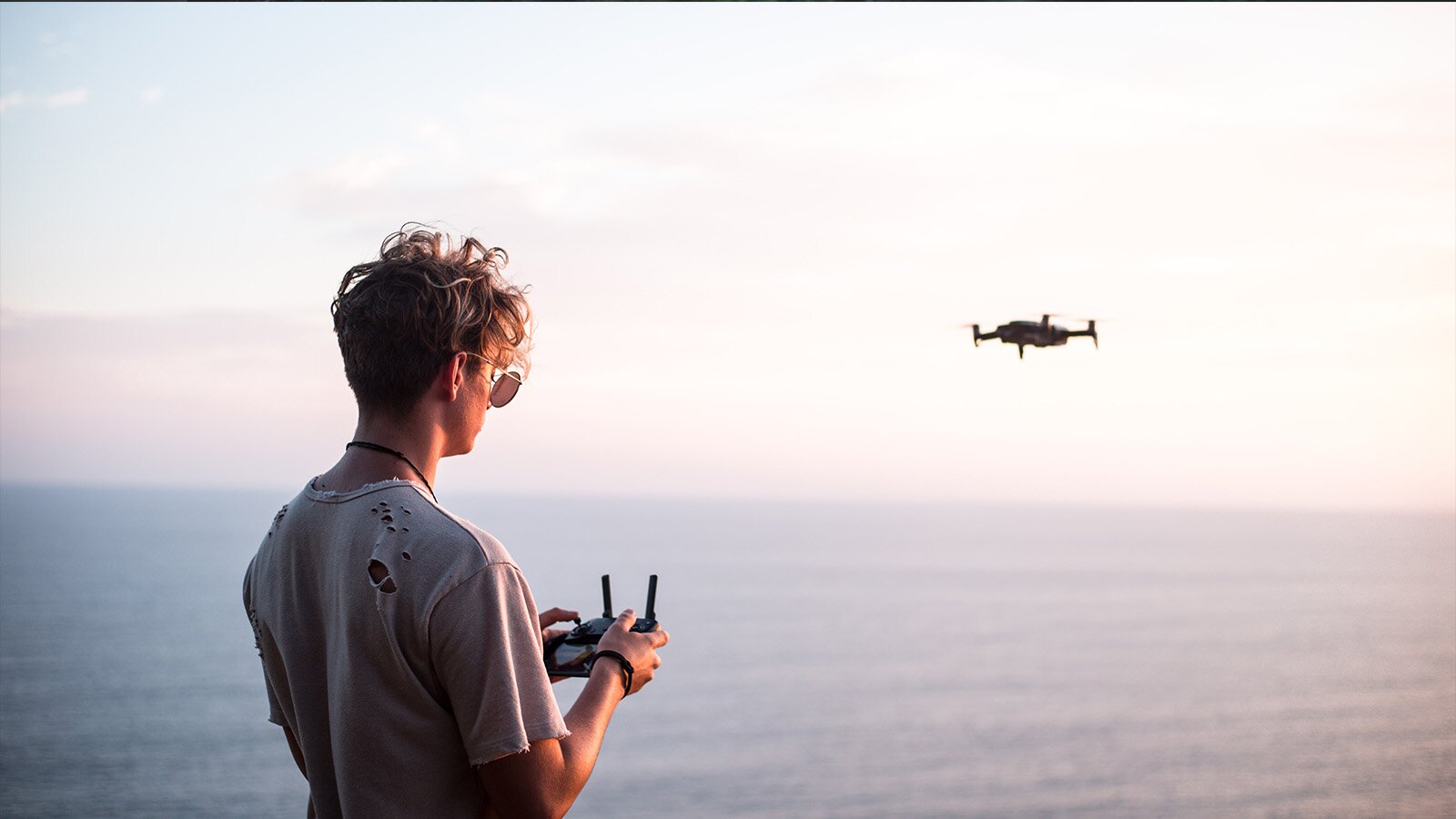 Zdjęcie przedstawia osobę stojącą nad morzem. Trzyma ona w dłoniach drążek sterowy kierujący dronem. 