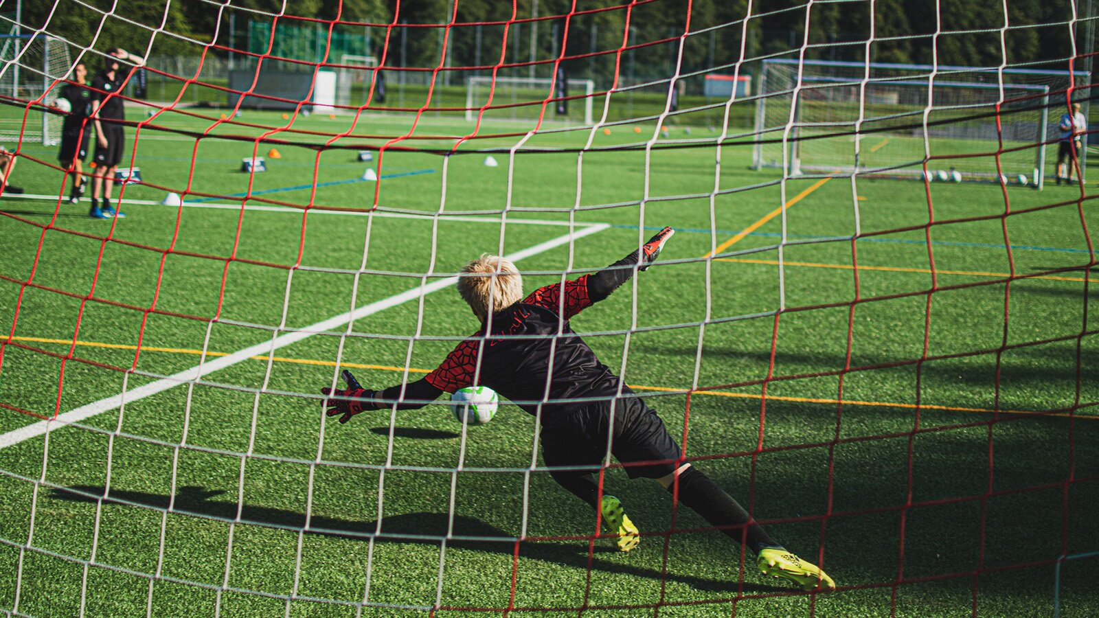 Zdjęcie przedstawia chłopaka w stroju bramkarze, który rzuca się na murawę, aby złapać znajdującą się przed nim piłkę. Ujęcie zostało zrobione zza bramki, więc widoczna jest siatka. W tle widoczna jest murawa boiska, inne bramki, porozrzucane po murawie piłki i dwie sylwetki dwóch osób stojących po lewej stronie.