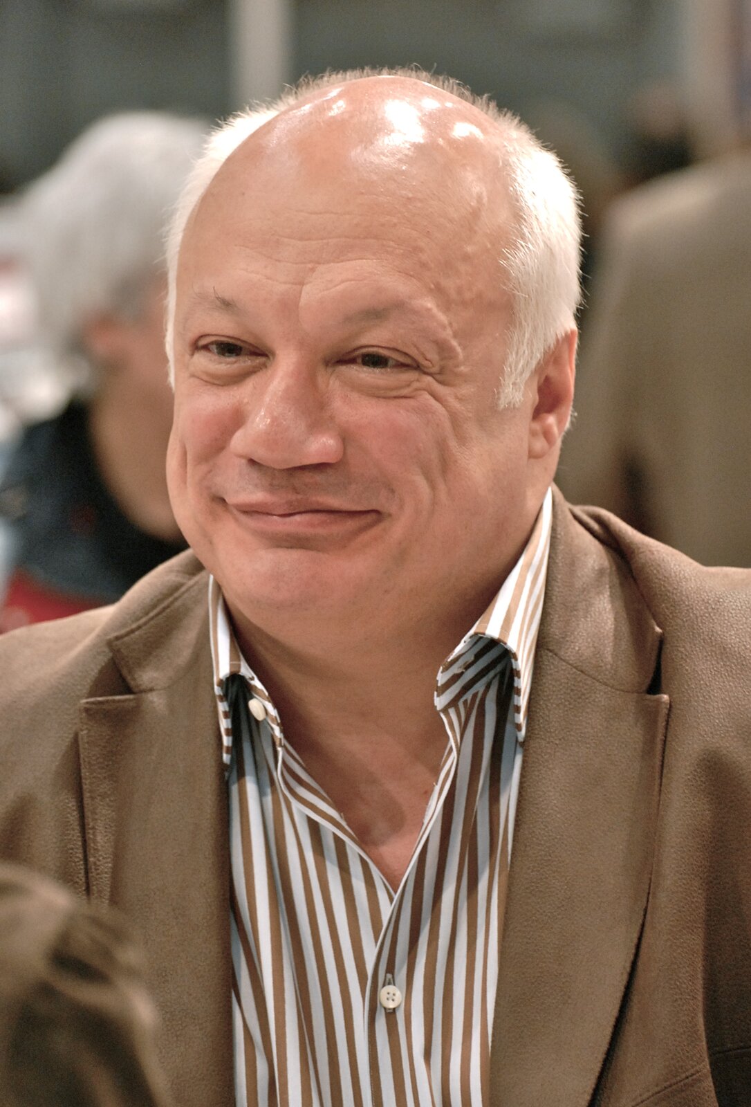 Fotografia barwna w układzie pionowym przedstawiająca portret, z lewego półprofilu, francuskiego pisarza Erica‑Emmanuela Schmitta. To dojrzały mężczyzna o egzotycznej urodzie ludów północnej Afryki. Pierwszy plan zajmuje jego głowa i wyłaniający się zza dolnej krawędzi kadru górny fragment klap beżowej marynarki oraz rozpiętej pod szyją koszuli w biało‑brązowe paski. Pełna, uśmiechnięta, jasna twarz pisarza zwrócona jest nieznacznie w lewo, w stronę osoby poza lewą krawędzią kadru. Widać tylko w dolnym narożniku kawałek jej ramienia. Sądząc po charakterystycznym dla damskiego odzienia marszczeniu w miejscu wszycia rękawa, jest to ramię kobiety. To do niej uśmiecha się pisarz, lekko mrużąc przy tym piwne oczy z wyraźnie opuchniętymi powiekami, nad którymi ledwie rysują się brwi. Nad nimi pokaźny wał czołowy podzielony pionową bruzdą skręcającą dolnym końcem w stronę lewego oka. Prawy kącik dość wydatnych i asymetrycznych ust uniósł się wyżej, a dolna warga tej ich strony cofnęła względem górnej, podczas gdy z lewej dolna warga jest wysunięta. Ogolona gładko twarz nosi jedynie nikły ślad odrastającego zarostu pod nosem, który jest krótki, prosty, ale mięsisty i szeroki. Pozbawiona włosów skóra na sklepieniu czaszki ponad wysokim czołem odbija światło niewidocznych na fotografii lamp. Łysinę okalają od tyłu włosy siwe i krótko przycięte, ale ich baczki sięgają połowy niewielkich uszu. W tle, w dużej nieostrości, w bliżej nieokreślonym wnętrzu, ludzkie postaci. Po lewej głowa posiwiałej kobiety, a po prawej plecy wysokiego mężczyzny w jasnej marynarce.