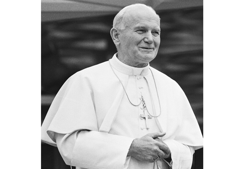 Czarno-biała fotografia przedstawiająca papieża Jana Pawła II. Ma na sobie białą szatę. Na jego szyi wisi duży krzyż. Papież się uśmiecha.