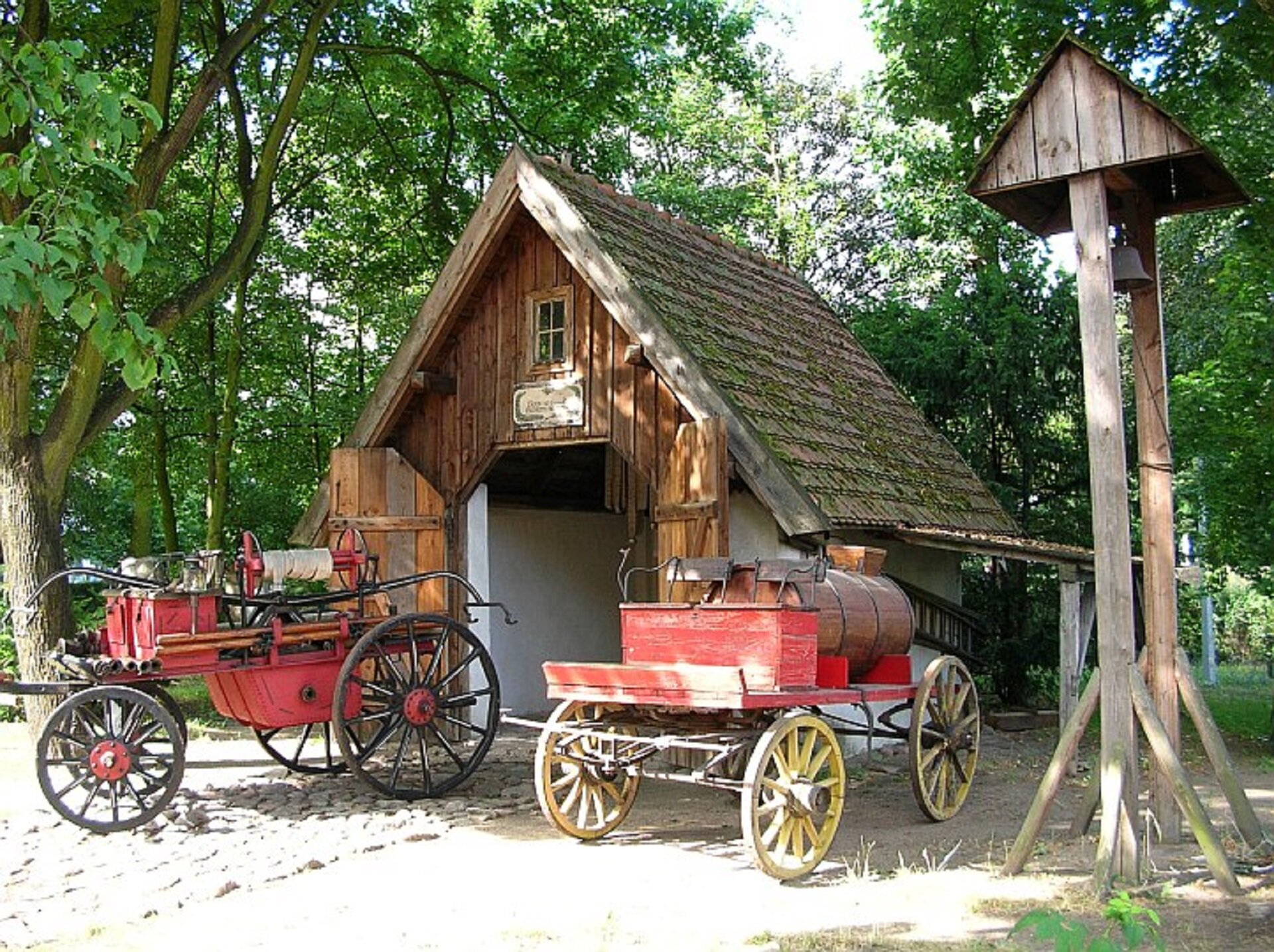Zdjęcie przedstawia starą remizę strażacką, będącą w posiadaniu Muzeum Etnograficznego w Toruniu. Budynek jest wykonany z drewna. Przed remizą stoją dwa stare, czerwone wozy strażackie. Z boku, po prawej stronie znajduje się niewielka, drewniana dzwonnica. W tle rosną liczne drzewa.