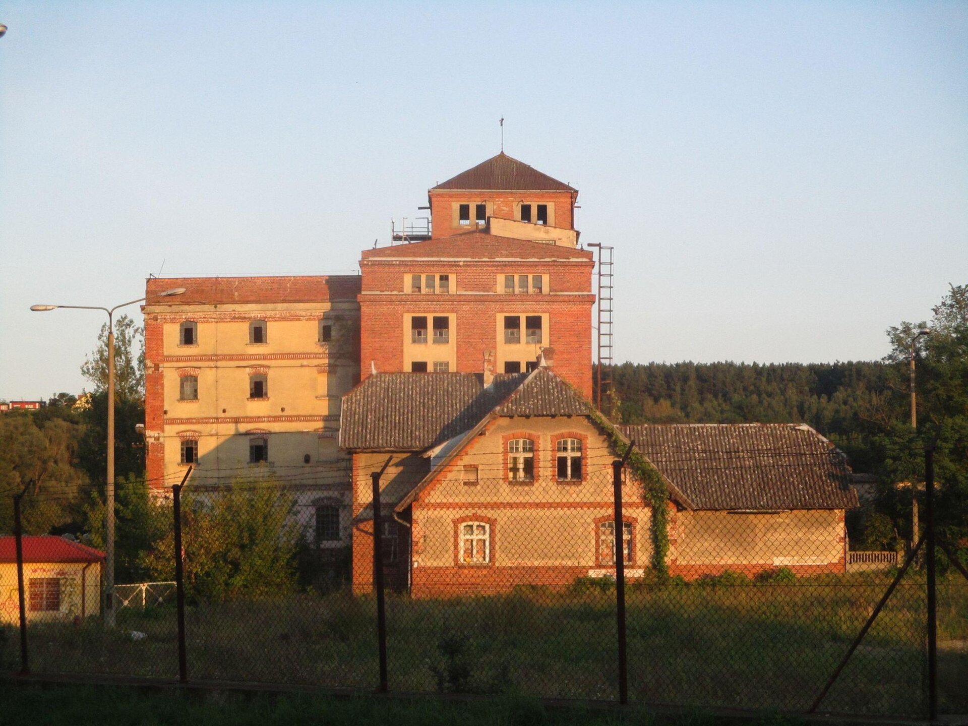 Zdjęcie przedstawia czteropiętrowy, pomarańczowy budynek. Przed nim stoi drugi, niższy, piętrowy, ze skośnym dachem. Oba budynki pokryte są czerwoną cegłą i kremowym tynkiem. Teren wokół zabudowań jest ogrodzony czarną siatką. Wokół budynków, a także za nimi, rosną zielone krzewy i drzewa. Niebo jest błękitne, bezchmurne. Zdjęcie zostało wykonane w świetle dziennym.