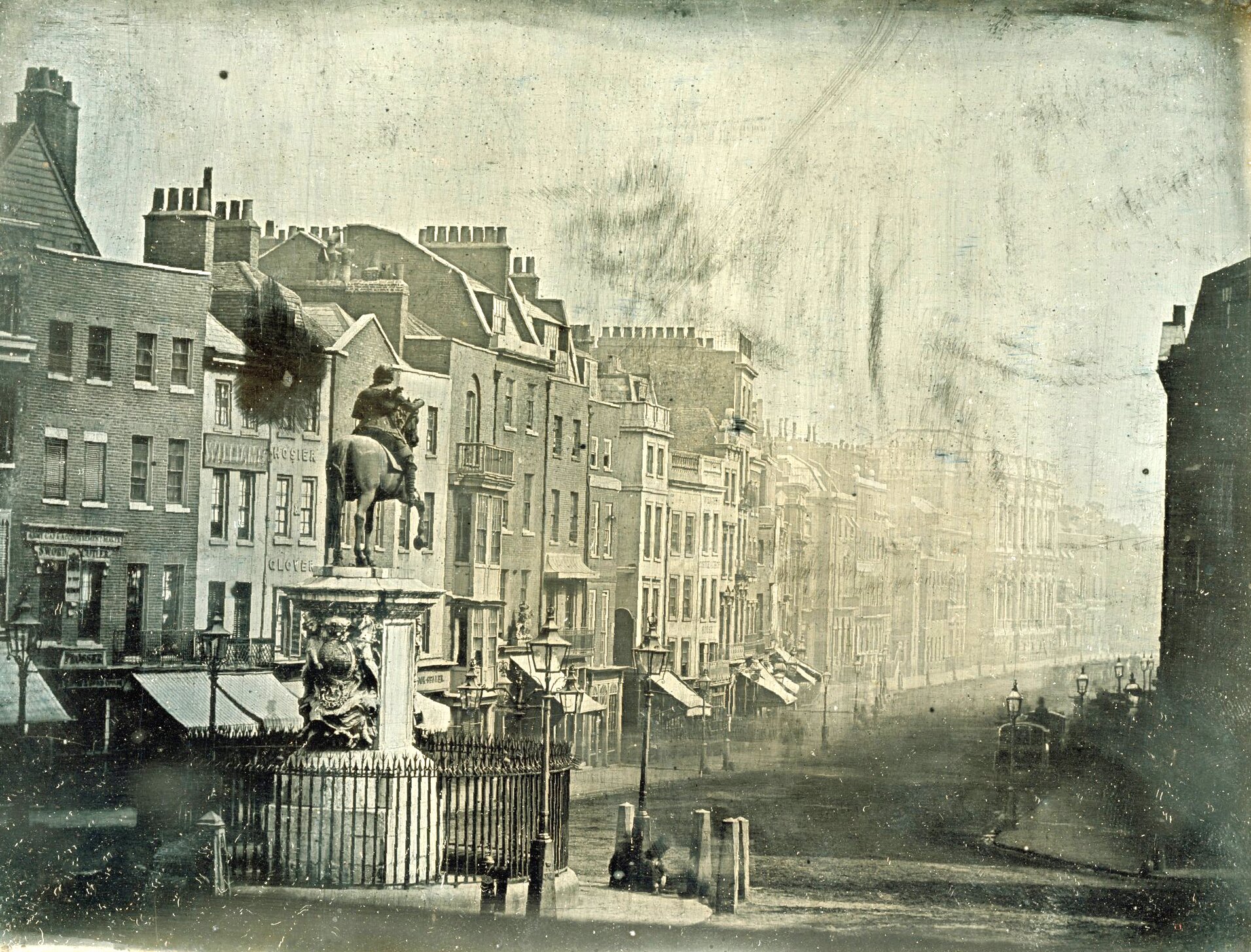 Stara czarnobiała fotografia fragmentu dziewiętnastowiecznego miasta. Pośrodku pomnik konny, przed nim aleja, po bokach niewysokie, wąskie budynki ustawione w szeregu.
