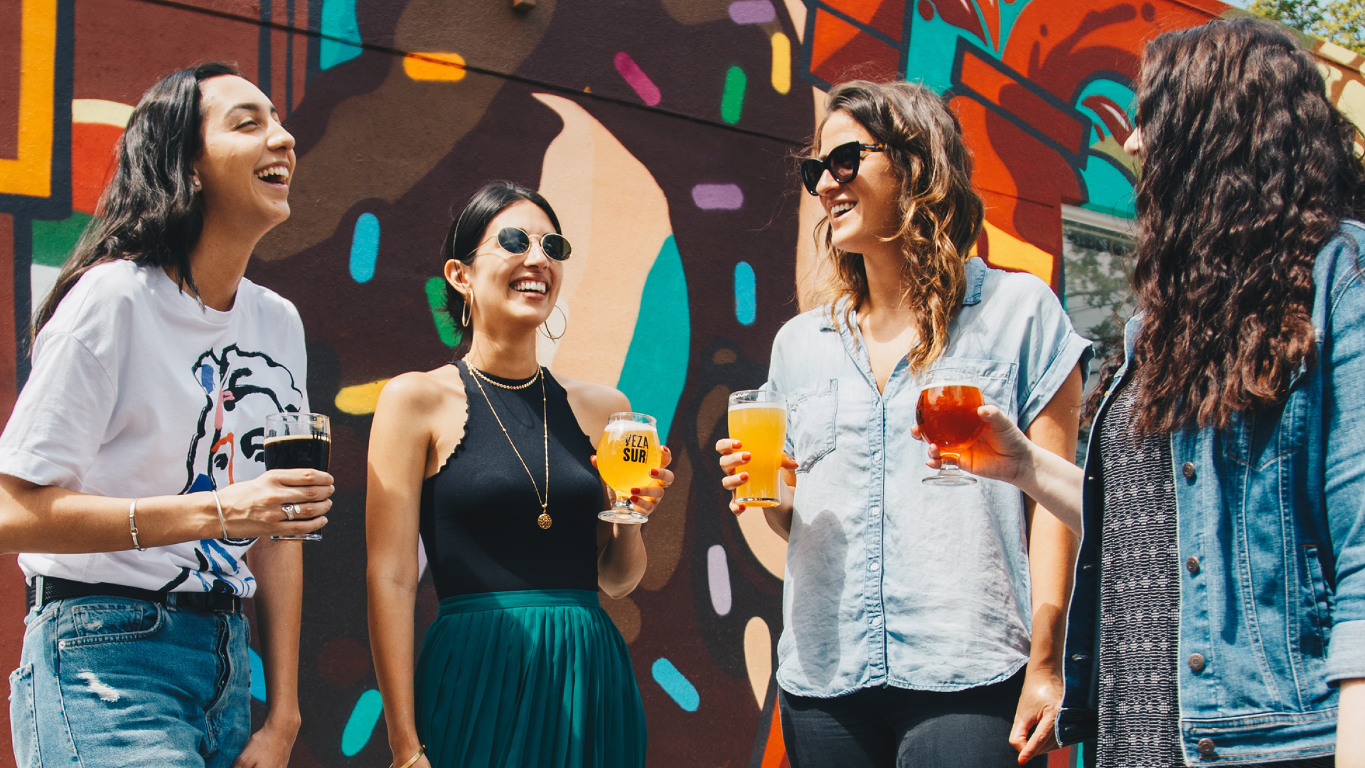 Zdjęcie przedstawia cztery uśmiechnięte dziewczyny. Stoją razem obok siebie, każda z nich trzyma w ręku jakiś napój w szklance.