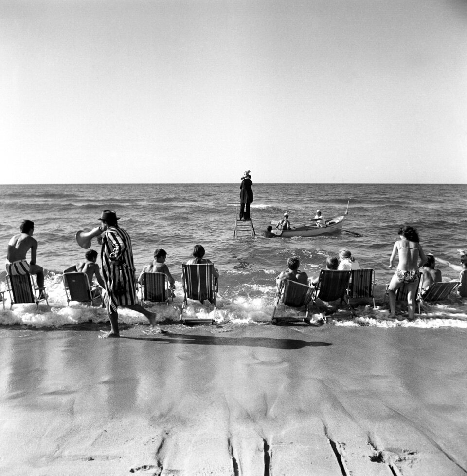Na starym czarno białym zdjęciu ukazana jest scena rozgrywająca się nad brzegiem morza. W płytkiej wodzie postawiono drabinę, na której stoi ubrany na czarno mężczyzna. Z prawej strony drabiny dryfuje łódka, w której siedzą dwaj mężczyźni trzymający w dłoniach wiosła. Trzeci mężczyzna stoi obok rufy łodzi. Na brzegu ustawiono osiem leżaków, w których siedzą ludzie. Za leżakami przechadza się mężczyzna ubrany w szlafrok w pionowe paski; w dłoni trzyma megafon. 