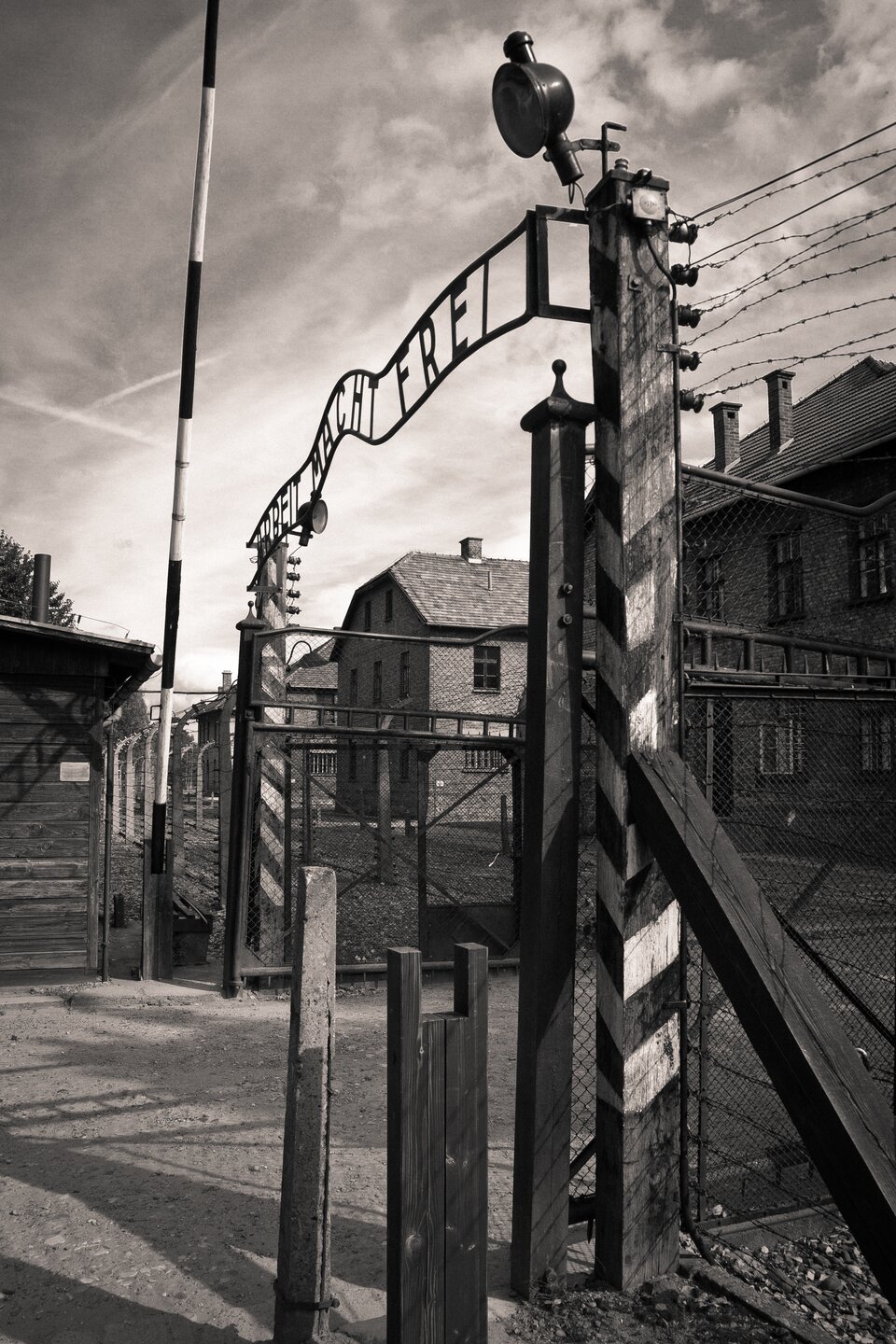 Fotografia przedstawiająca bramę wejściową z napisemArbeit macht frei . Przed bramą otwarty szlaban, nad siatką ogrodzenia umieszczone druty kolczaste. Na drugim planie dwukondygnacyjne baraki.