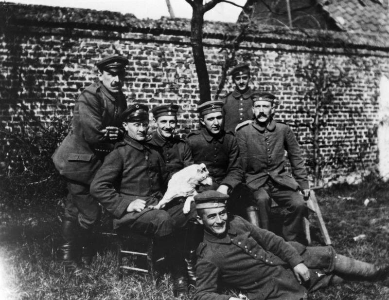 Kliknij, aby powiększyć Zdjęcie przedstawia siedmiu żołnierzy. Czterech z nich siedzi na krzesłach. Jeden z nich trzyma na kolanach psa. Dwoje żołnierzy stoi, zaś jeden leży przed tymi co siedzą na krzesłach. Pierwszy od prawej siedzący żołnierz to Hitler z charakterystycznymi szerokimi wąsami. Tło zdjęcia stanowi mur z cegieł. Na ziemi widać trawę.