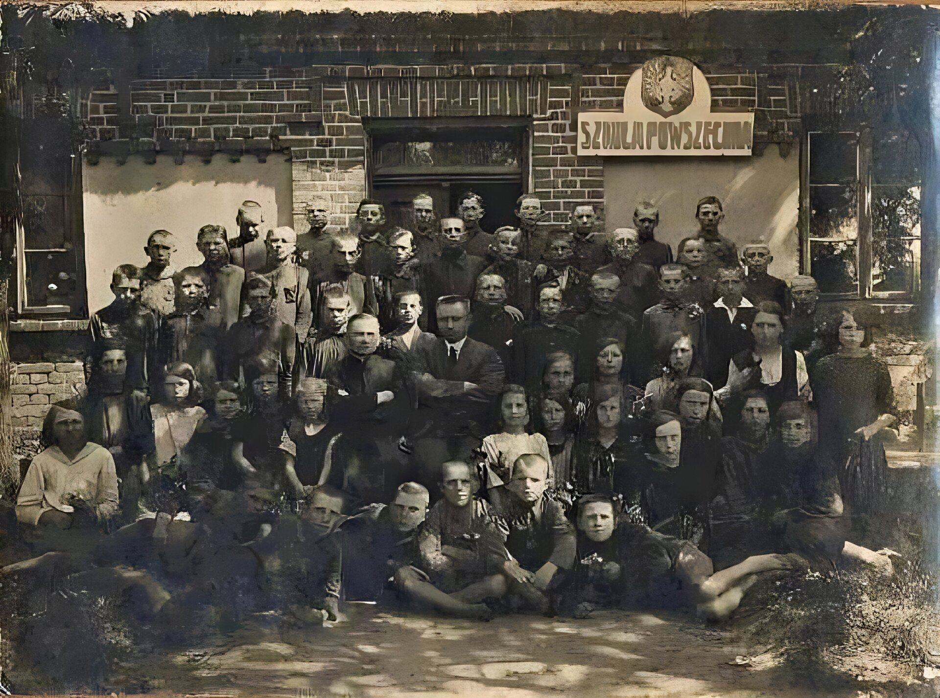 Uczniowie przed budynkiem szkoły w Czernikowie - rok 1919.  Zdjęcie w odcieniach szarości.  Grupa około sześćdziesięciu dzieci (chłopców i dziewczynek) pozuje do zdjęcia. Między dziećmi siedzi dwóch mężczyzn.  W tle ceglany budynek. Na nim godło polski i napis - "Szkoła podstawowa".