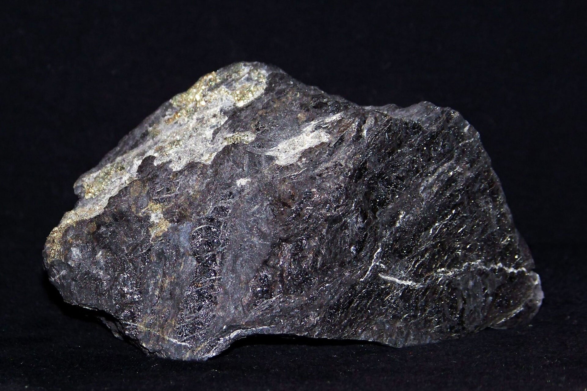 Fotografia przedstawia czarną bryłkę węgla kamiennego. Skała osadowa pochodzenia roślinnego. W przedstawionej bryle widać widoczne warstwy, zagłębienia pęknięcia, nierówności. Bryła ma dość gładkie krawędzie, ociosane w naturalny sposób.