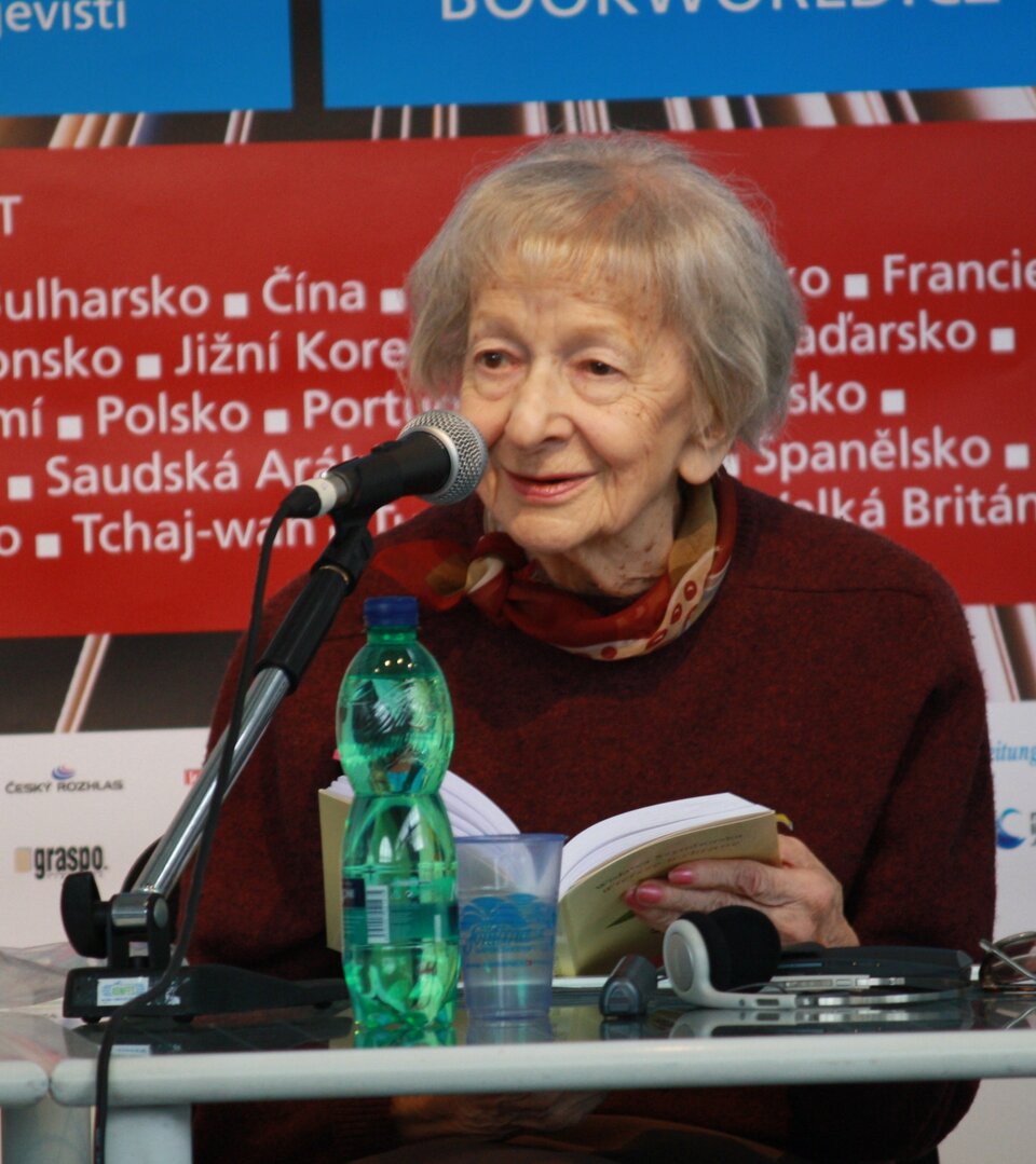 Zdjęcie przedstawia starszą, siwowłosą kobietę. Jej twarz jest szczupła, widoczne są na niej zmarszczki. Kobieta uśmiecha się. Siedzi przy stole, przed mikrofonem. Trzyma w dłoniach otwartą książkę.