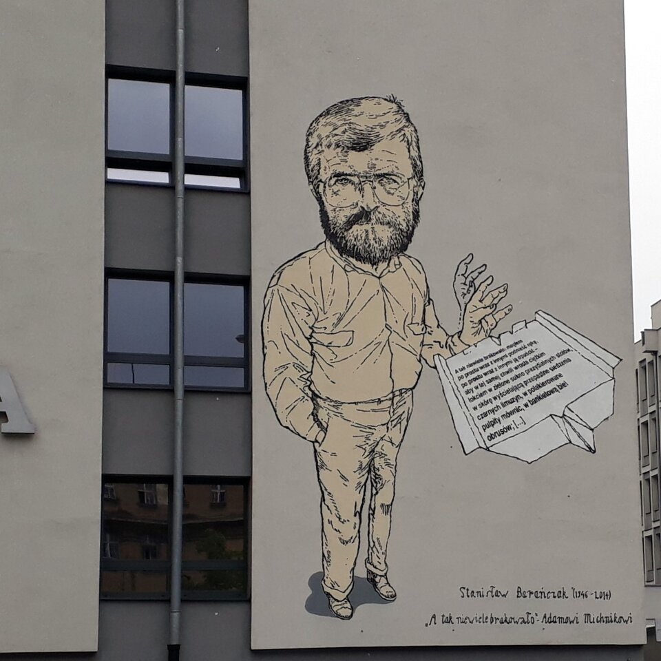 Zdjęcie przedstawia mural na ścianie dużego budynku. Namalowany jest dojrzały, brodaty mężczyzna. Z lewej ręki wypuszcza on złożoną w kształt samolotu, zapisaną kartkę papieru.
