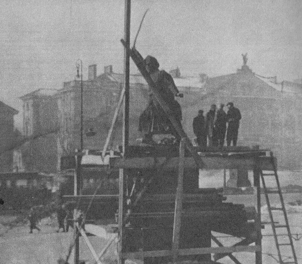 Zdjęcie przedstawia pomnik usytuowany na wysokim cokole, wokół którego stoją rusztowania. Na rusztowaniu stoi kilka osób. W tle widać kamienice.  