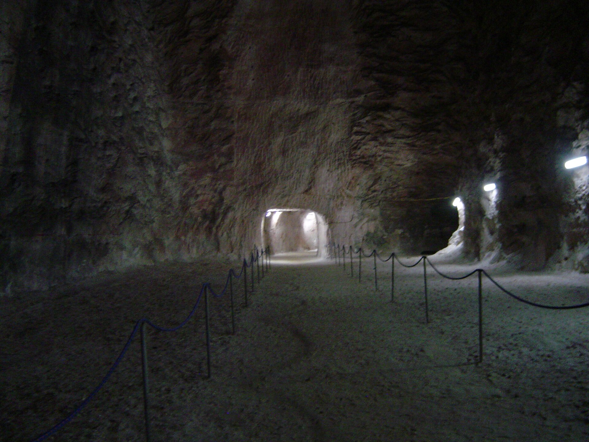 Zdjęcie ukazuje jedną z komór w kopalni soli w Kłodawie. Komora ma skalne, wysokie sklepienie przypominające sufit jaskini. Dno komory jest płaskie i białawe. W tle widoczny podświetlony tunel, którym zwiedzający przechodzą do kolejnych miejsc kopalni.