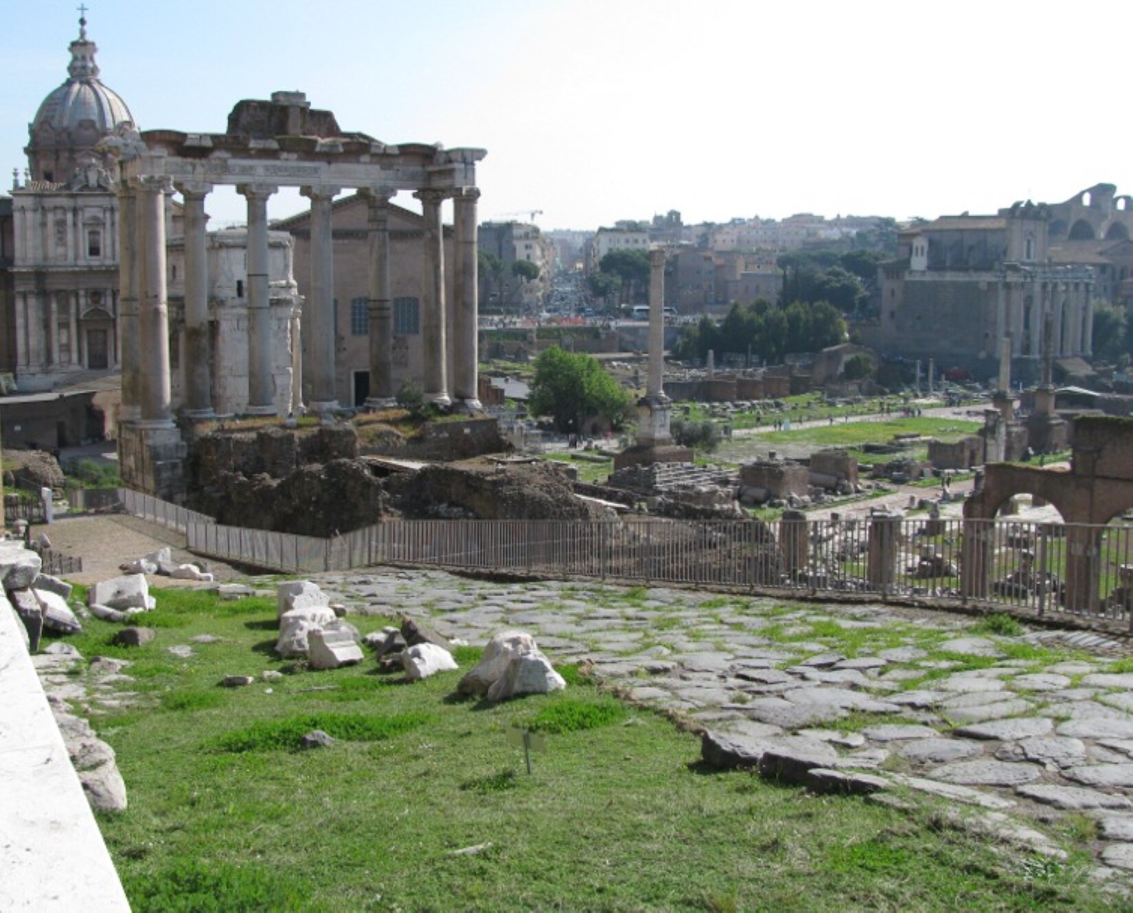 Fotografia przedstawia kamienistą drogą biegnącą ku górze. Kamienie na drodze są ścięte na podobnej wysokości i niemal nie wystają nad powierzchnię. Wzdłuż drogi ustawiono barierkę zabezpieczającą turystów przed upadkiem z wysokości. W tle ruiny oraz pozostałości po budowlach starorzymskich. Fotografia została wykonana w jasny i bezchmurny dzień.