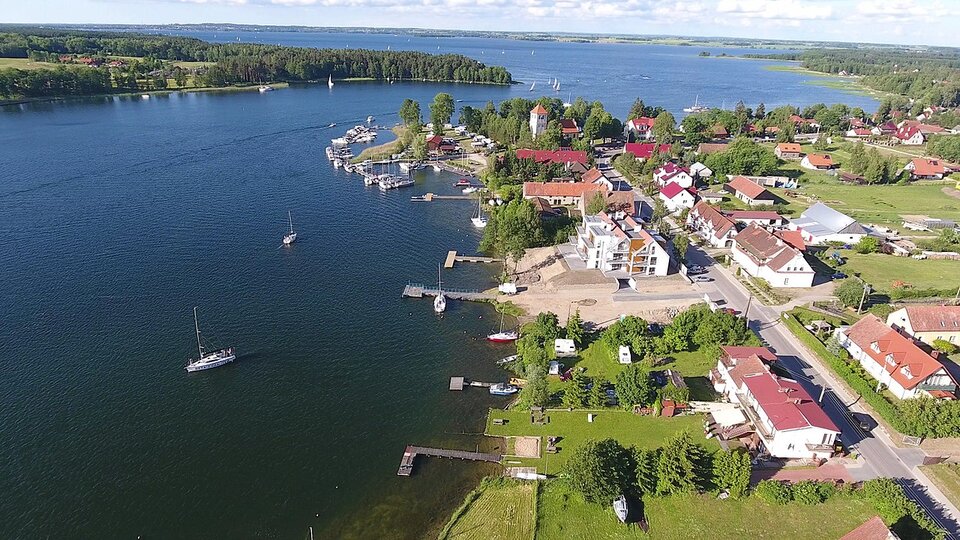Na zdjęciu jest jezioro z zabudowaniami nad jego brzegiem. Jezioro zwęża się w jednym miejscu i ponownie jest szerokie. 