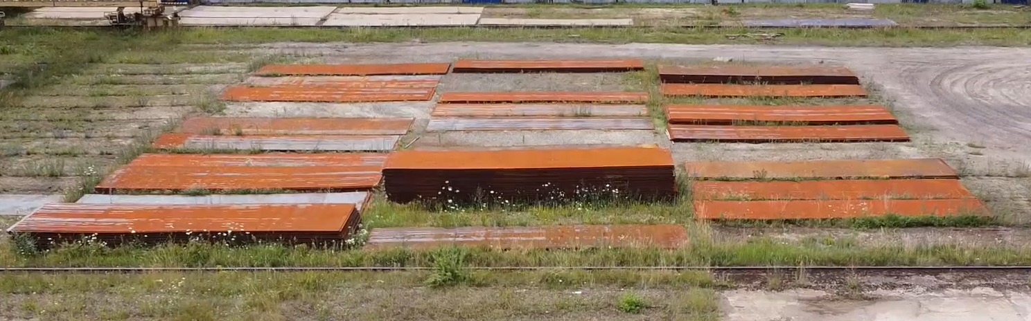 Zdjęcie przedstawia skorodowane blachy. Leżą one w stertach, jedna na drugiej, na placu za budynkiem stoczni. Pokryte są rdzawym nalotem.