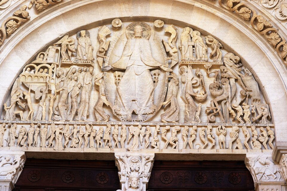 Romański tympanon portalu katedry: w centrum monumentalna postać Chrystusa w majestacie z rozłożonymi ramionami, po bokach aniołowie oraz sceny Sądu Ostatecznego z duszami zbawionymi i potępionymi; poniżej pas drobnych postaci w procesji, całość wykuta w jasnym kamieniu i ujęta półkolistym łukiem.