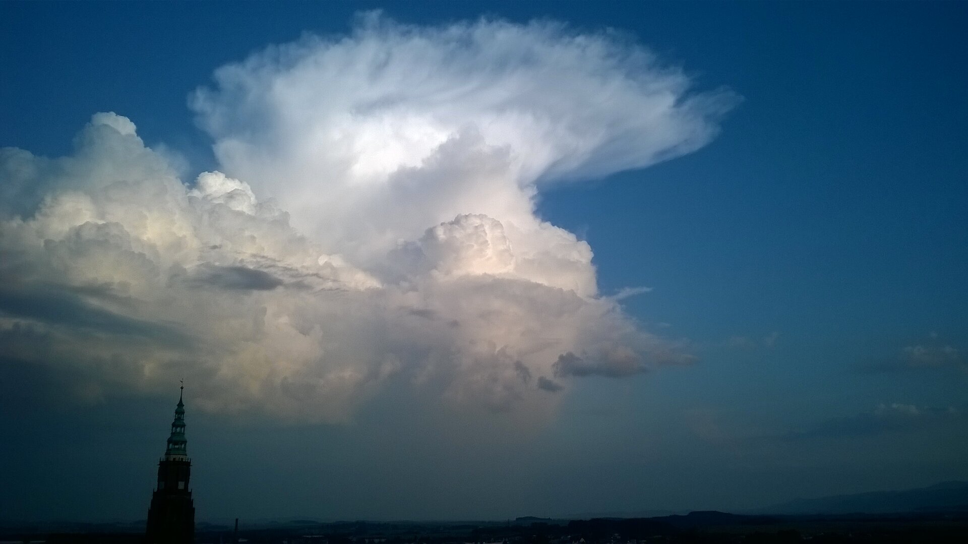 Fotografia przedstawia chmury typu cumulonimbus. Jest ona rozległa, biała, kłębiasta o nieregularnym kształcie, znajduje się nad wysoką, spiczastą wieżą budynku, na tle ciemno‑niebieskiego nieba.