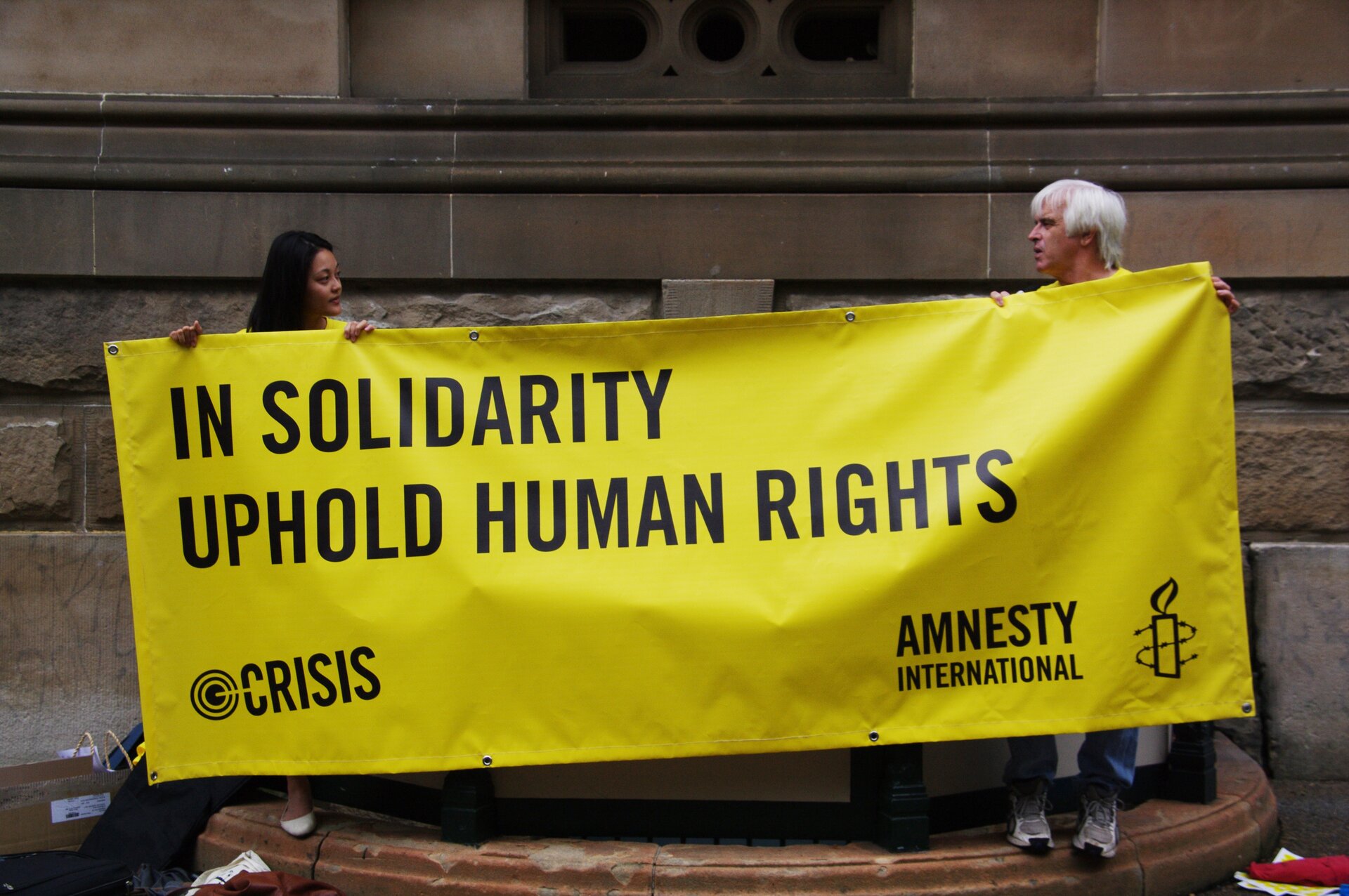 Fotografia barwna przedstawiająca dwójkę ludzi trzymających banner Amnesty International. Główny napis na bannerze głosi "In Solidarity Uphold Human Rights". W lewym dolnym rogu znajduje się napis "crisis" a w prawym - logo i nazwa organizacji Amnesty International.