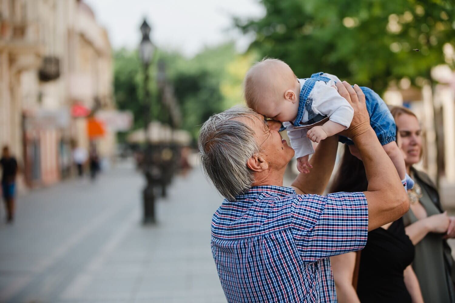 Zdjęcie przedstawia starszego mężczyznę z małym dzieckiem na miejskim deptaku w letni dzień. Zajmują oni prawą stronę kadru. Mężczyzna stoi profilem w stronę obiektywu. Ma dłuższe szpakowate włosy. Ubrany jest w koszulę z krótkim rękawem w kolorową kratkę. Dziecko ma około pół roku. Ubrane jest w dżinsowe ogrodniczki i białą koszulkę, nie ma włosów. Mężczyzna trzyma dziecko w górze obiema rękami i dotyka nosem i czołem nosa i czoła dziecka. Mężczyzna uśmiecha się. W tle widoczna jest kobieta i deptak miejski z latarniami, chodnikiem, drzewami i kamienicami.