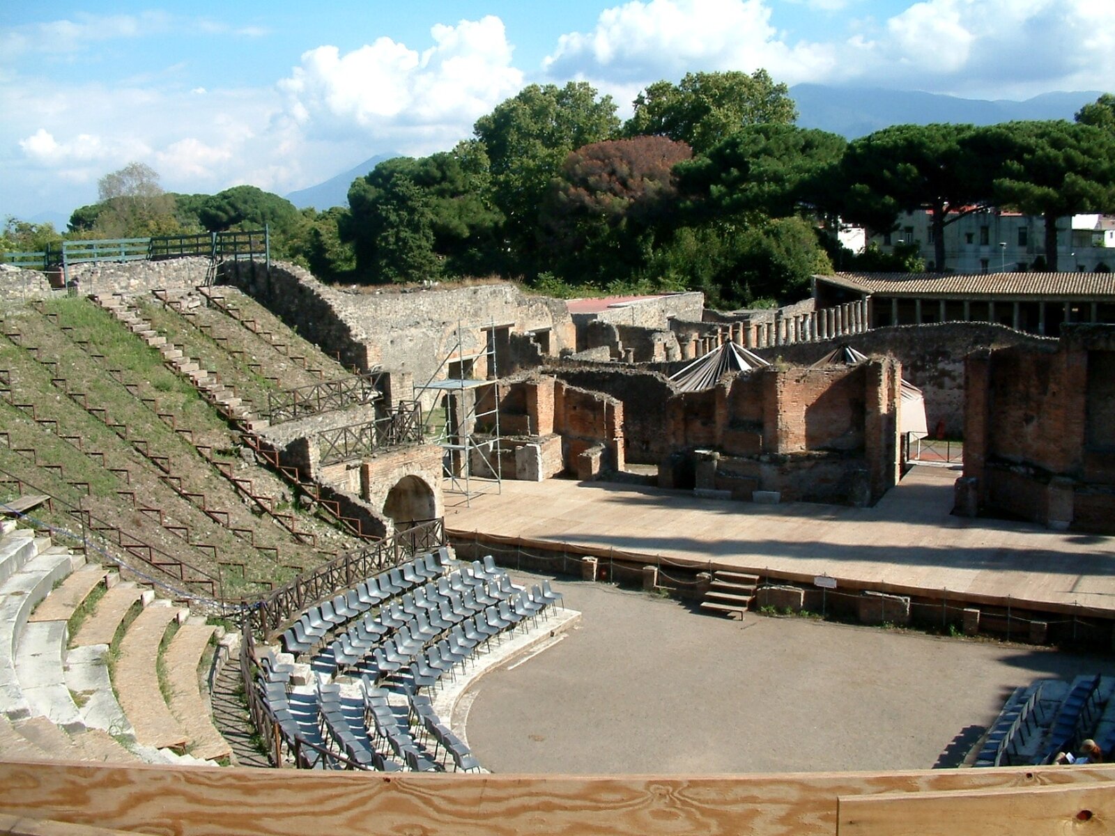 Na zdjęciu teatr na świeżym powietrzu, amfiteatr wbudowany w naturalne zagłębienie terenu. Widownia znajduje się po lewej stronie zdjęcia w wyższych rzędach z płyt kamiennych. Cztery pierwsze rzędy tworzą szaro‑stalowe, podobne do współczesnych krzesła. Widownia ma kształt półkola, w którego środku znajduje się orchestra – półokrągły plac między sceną a widownią z szarą podłogą, która przypomina klepisko. Scena znajduje się po prawej stronie zdjęcia, niemal w centrum. Ma kolor jasnobeżowy, kamienny. Prowadzą na nią czterostopniowe schodki. Na tyłach sceny widoczne są ściany i korytarze z cegły. Część amfiteatru i widowni pokrywa metalowe rusztowanie.
W kolejnym planie dalsza zabudowa teatru oraz gęsta roślinność ¬¬– zielone drzewa. W tle góry i niebieskie niebo pokryte białymi chmurami.