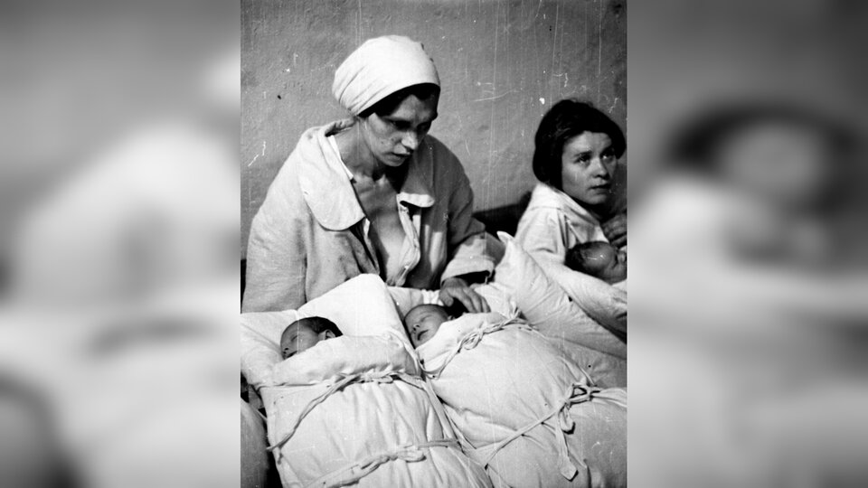 Zdjęcie przedstawia kilkoro niemowląt owiniętych w białe beciki. Nad dziećmi siedzą i czuwają dwie kobiety w białych ubraniach.