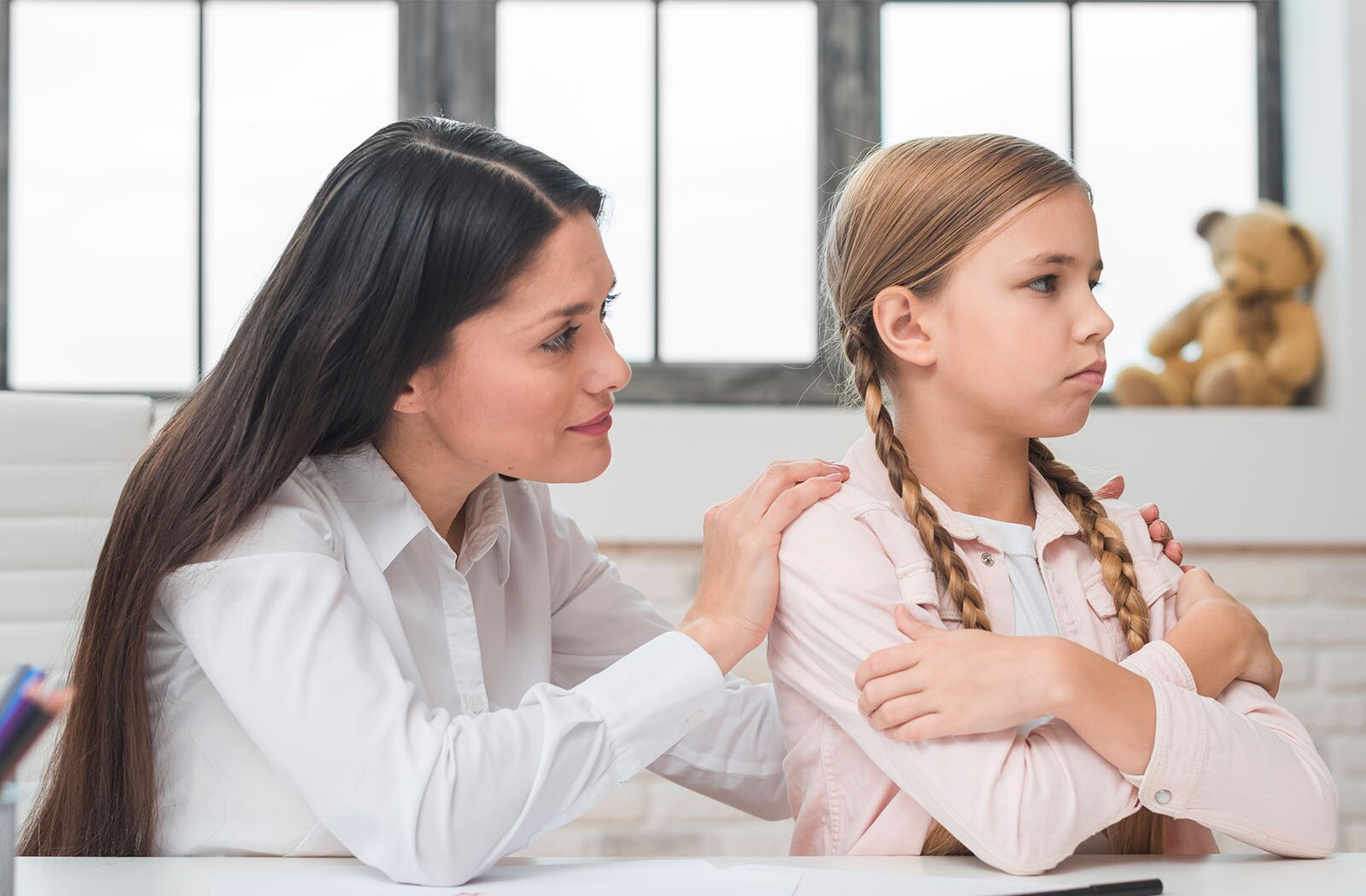 Zdjęcie przedstawia siedzącą przy stole dziewczynkę w wieku około dziesięciu lat. Dziewczynka obejmuje się rękoma i ma smutną minę. Za dziewczynką siedzi kobieta w młodym wieku, która łagodnym wzrokiem obserwuje dziewczynkę i trzyma dłoń na jej ramieniu.